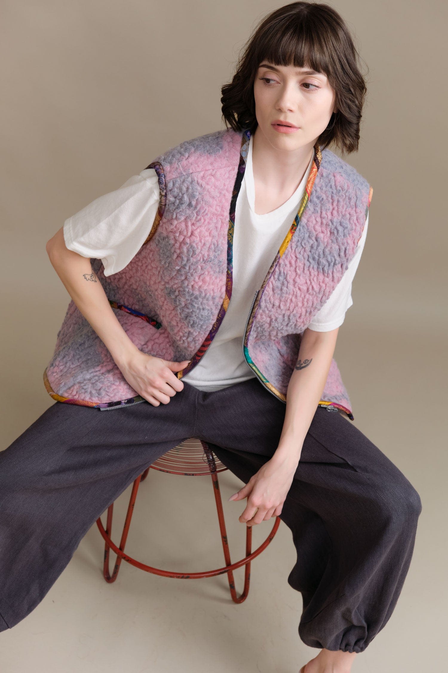 Person wearing a colorful Anntian vest sitting on a stool against a beige background