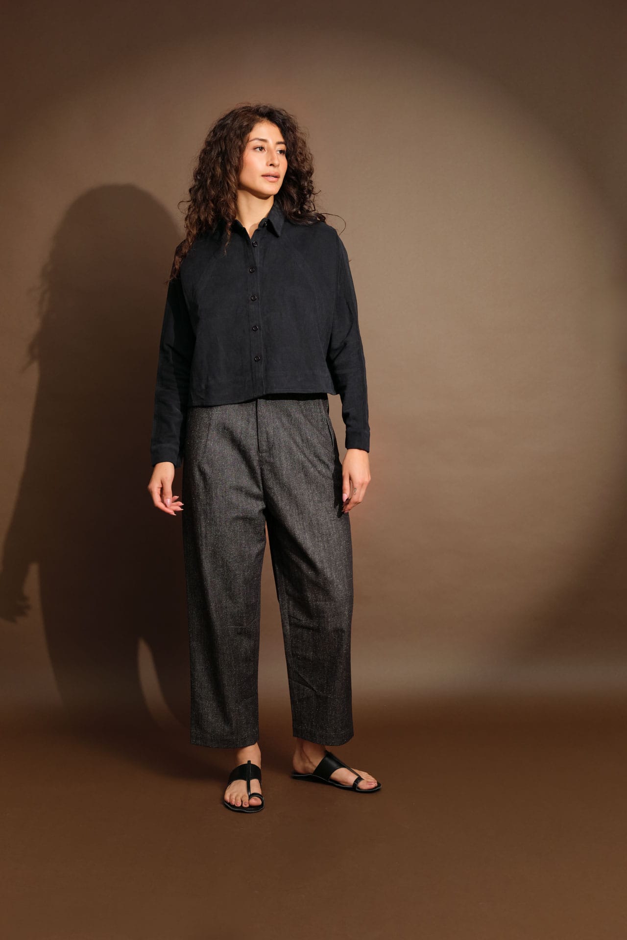 A model stands against a warm brown studio backdrop wearing a 7115 by Szeki dark navy long-sleeve button-up shirt, dark gray wide-leg trousers, and black slide sandals.