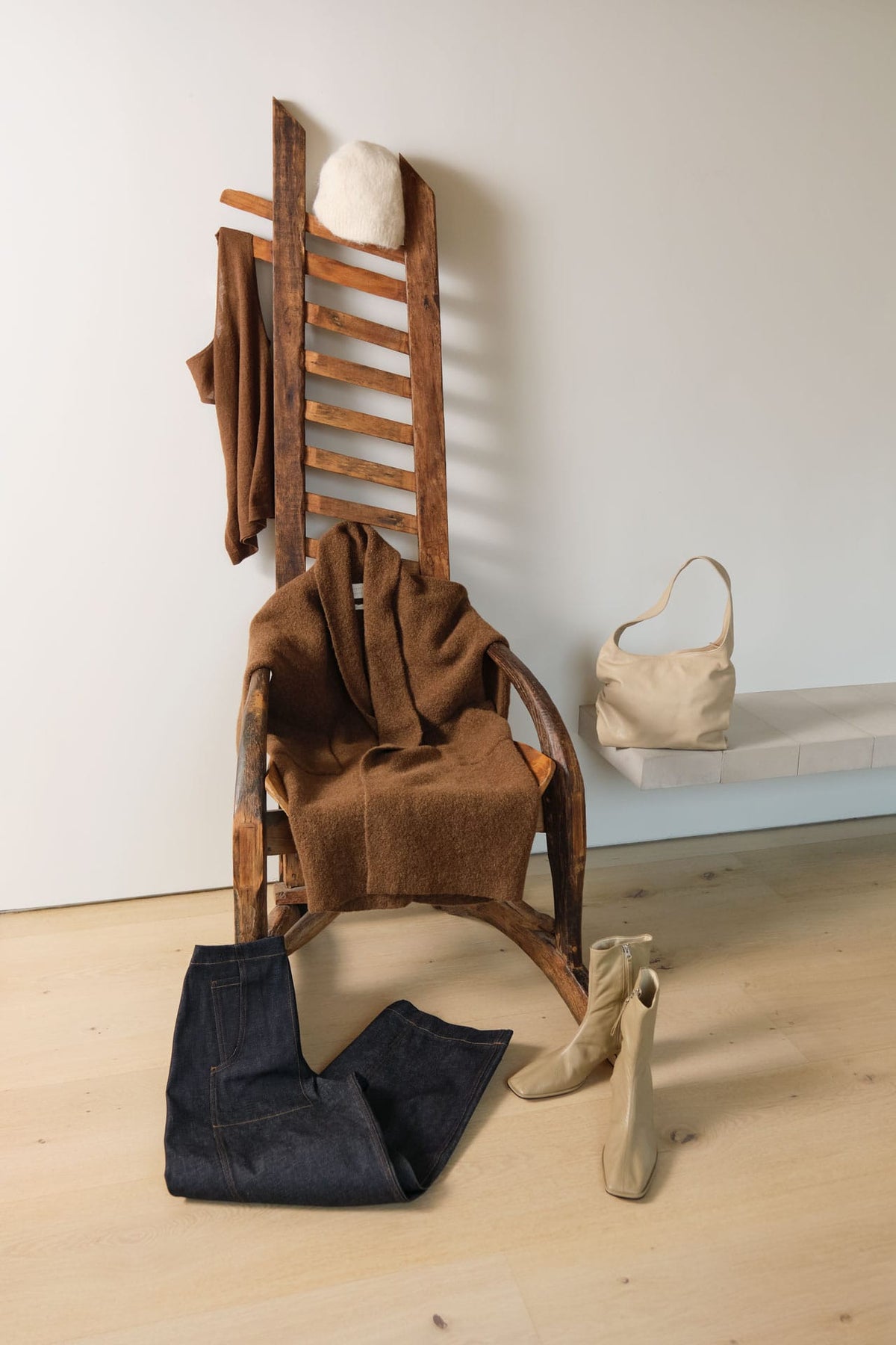 A styled fashion scene in a minimalist room with a wooden chair, draped brown knit clothing, folded dark blue jeans, beige ankle boots, a cream handbag on a bench, and a white hat, against a plain light wall and pale wood floor.