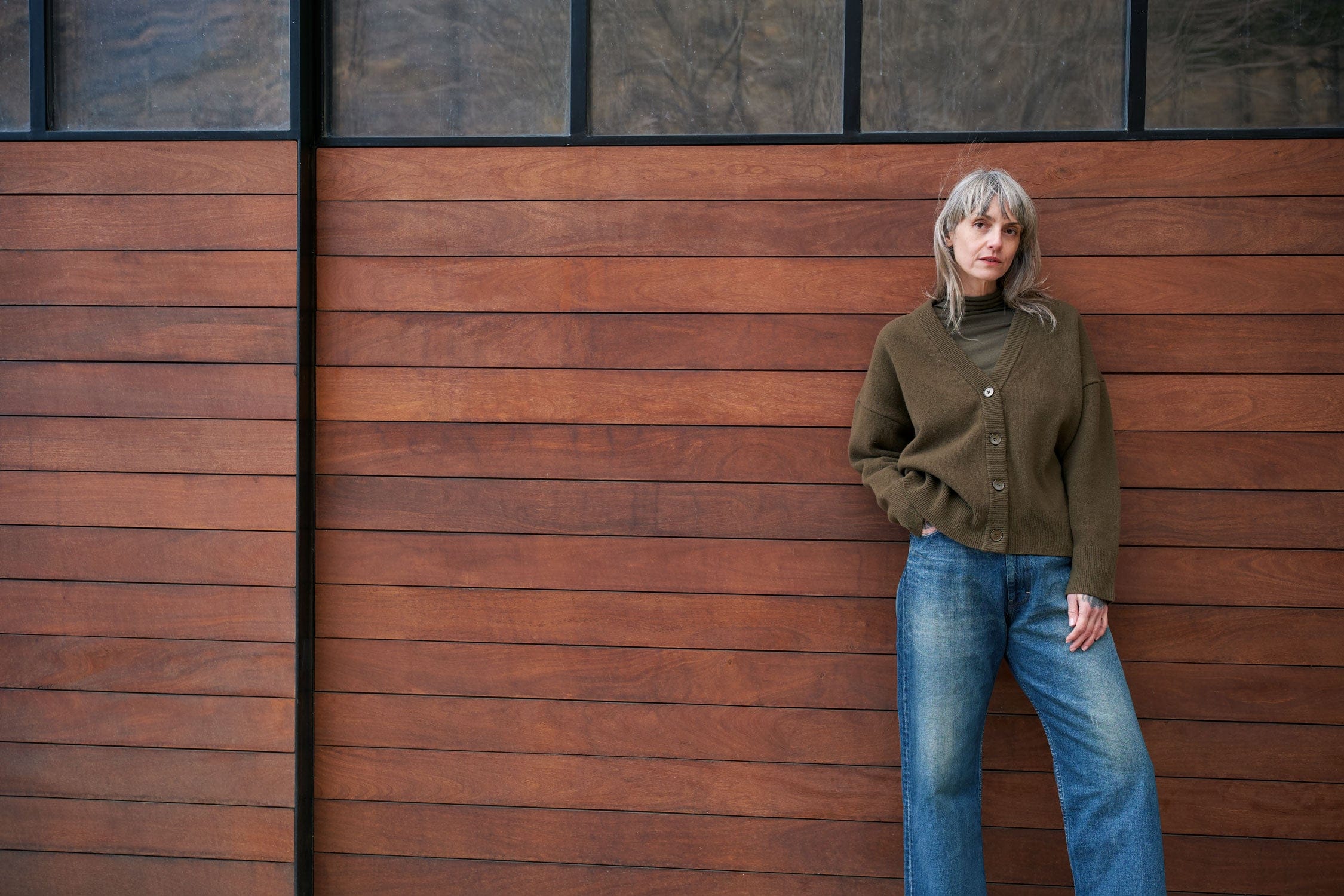 Person wearing Christian Wijnants and Chimala jeans leaning against a wooden wall with a modern building in the background