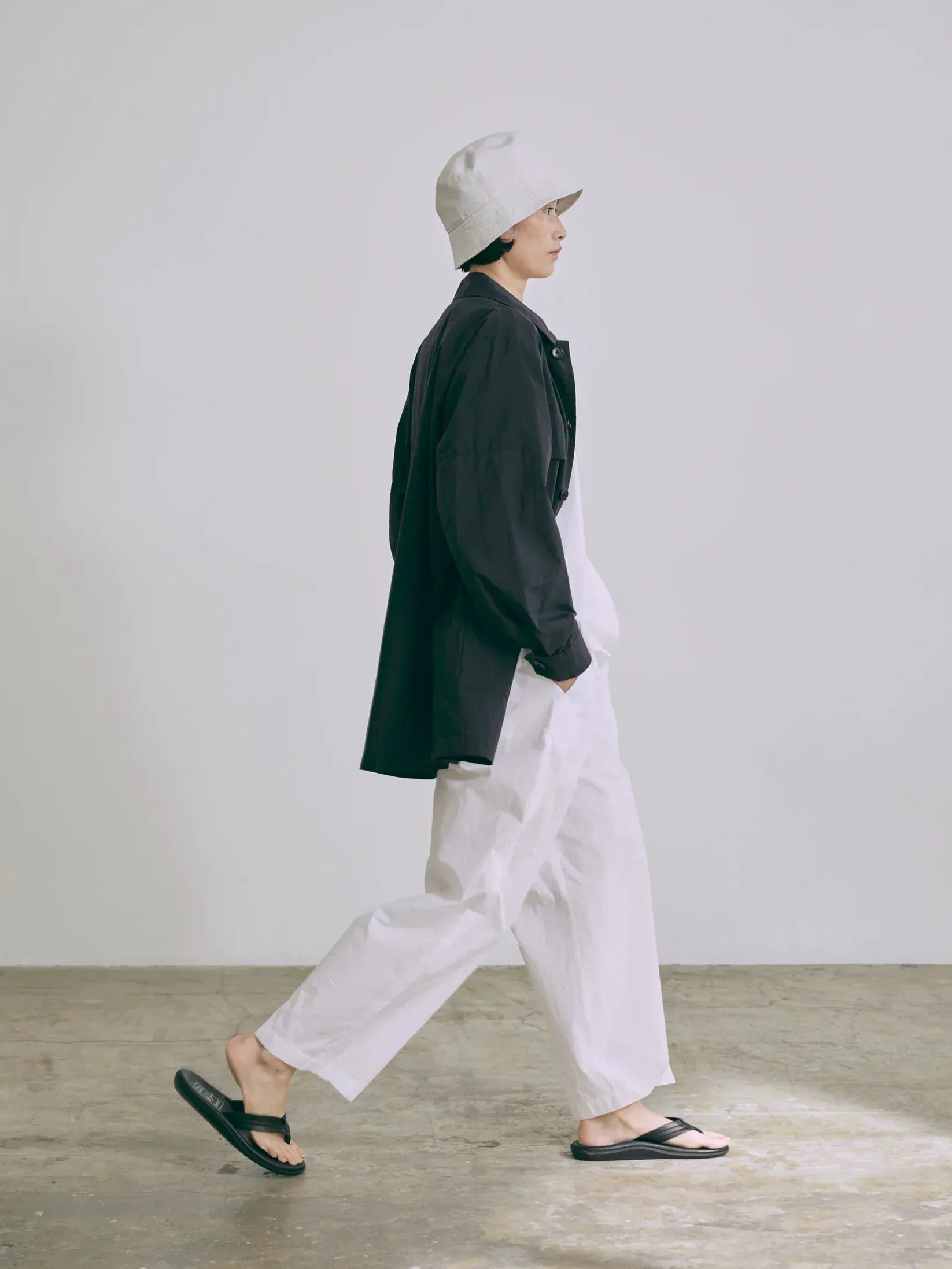 Person wearing a Nicholson & Nicholson black coat and white pants walking on a wooden floor with a plain background