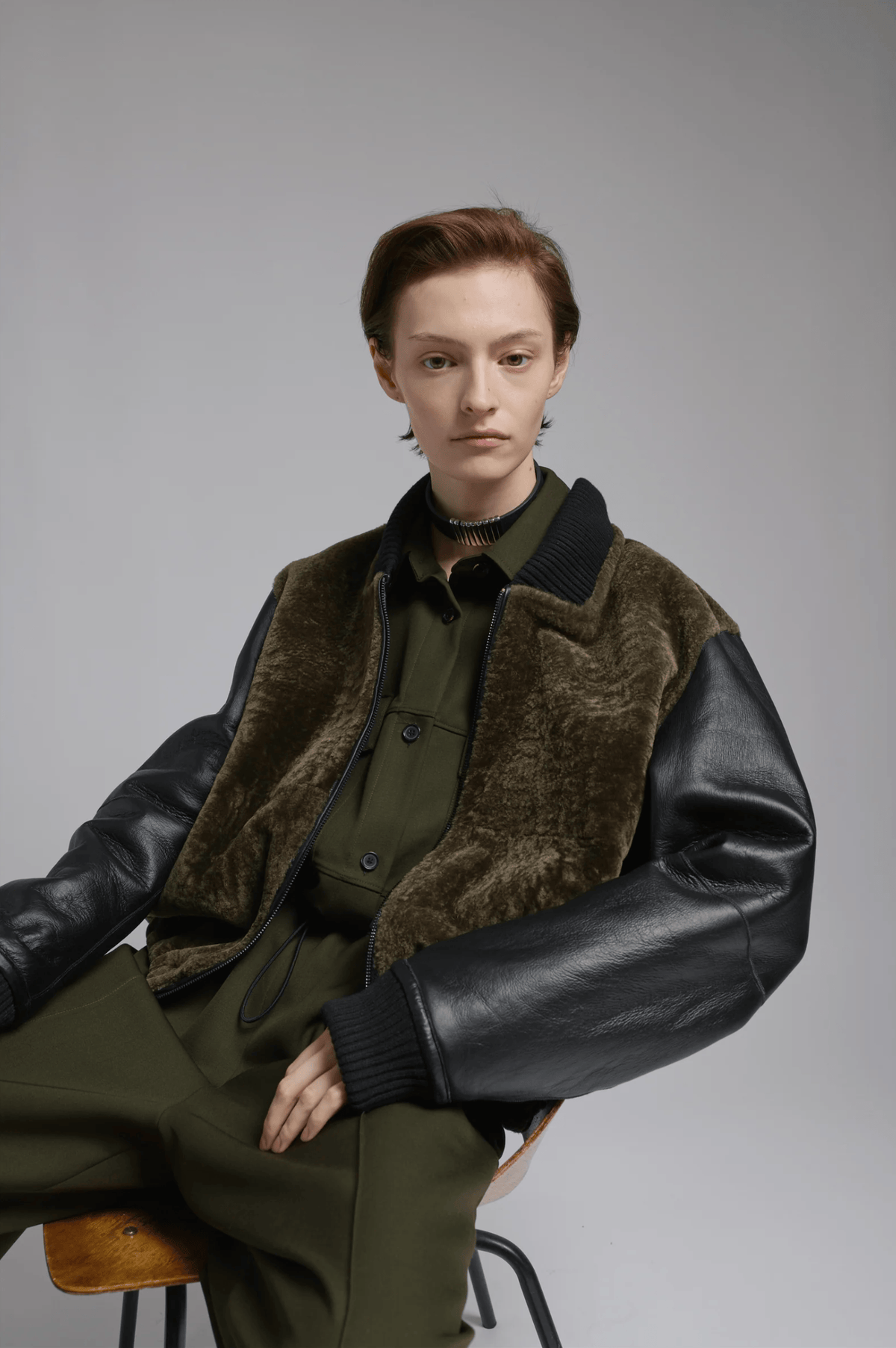 A model wearing a Rachel Comey dark olive green uniform-style jacket with contrasting black leather sleeves and a brown faux-fur collar, seated on a stool against a neutral gray background.