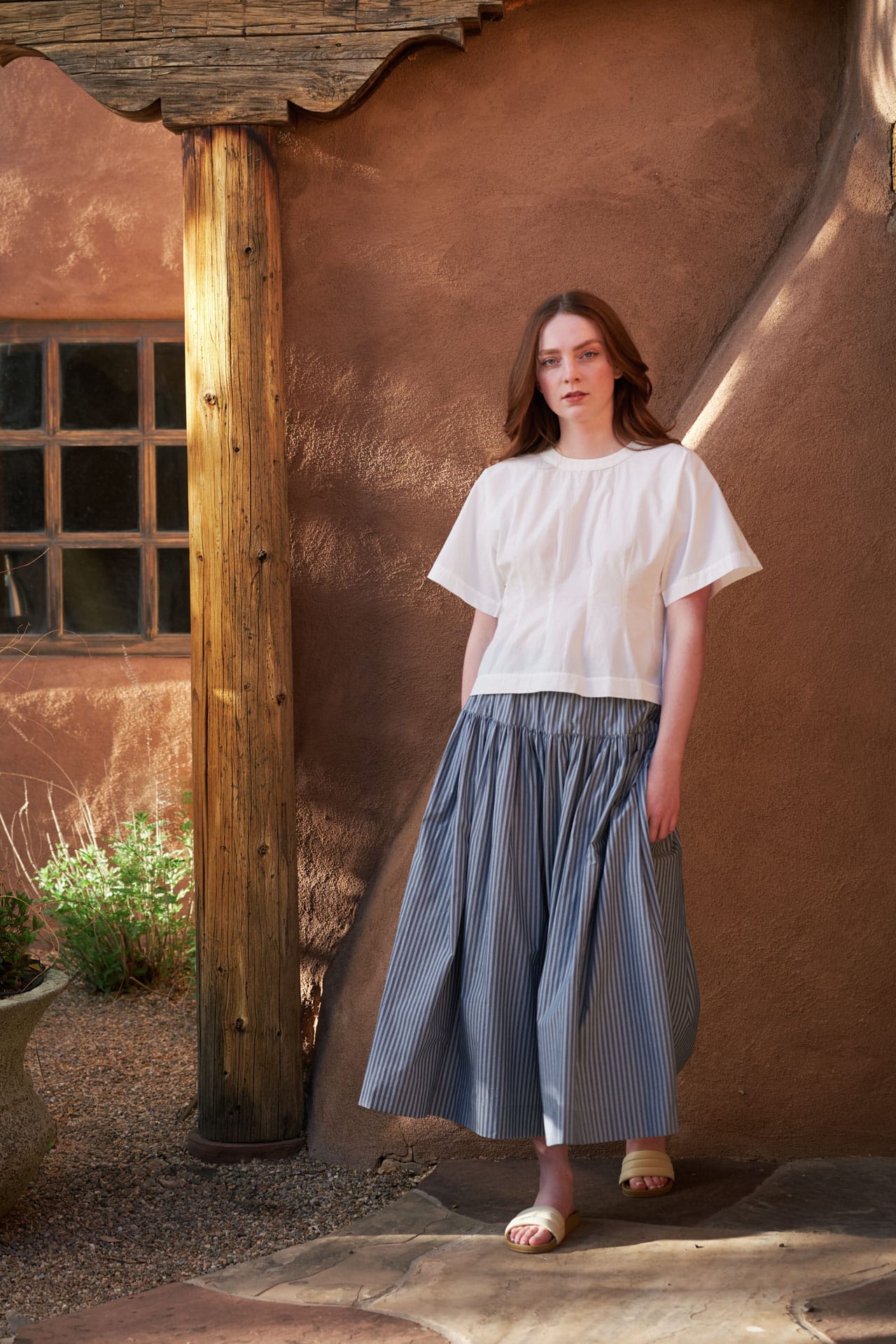 A woman in a Mirth white top and A'Court striped blue skirt standing in front of a rustic building with a wooden door.