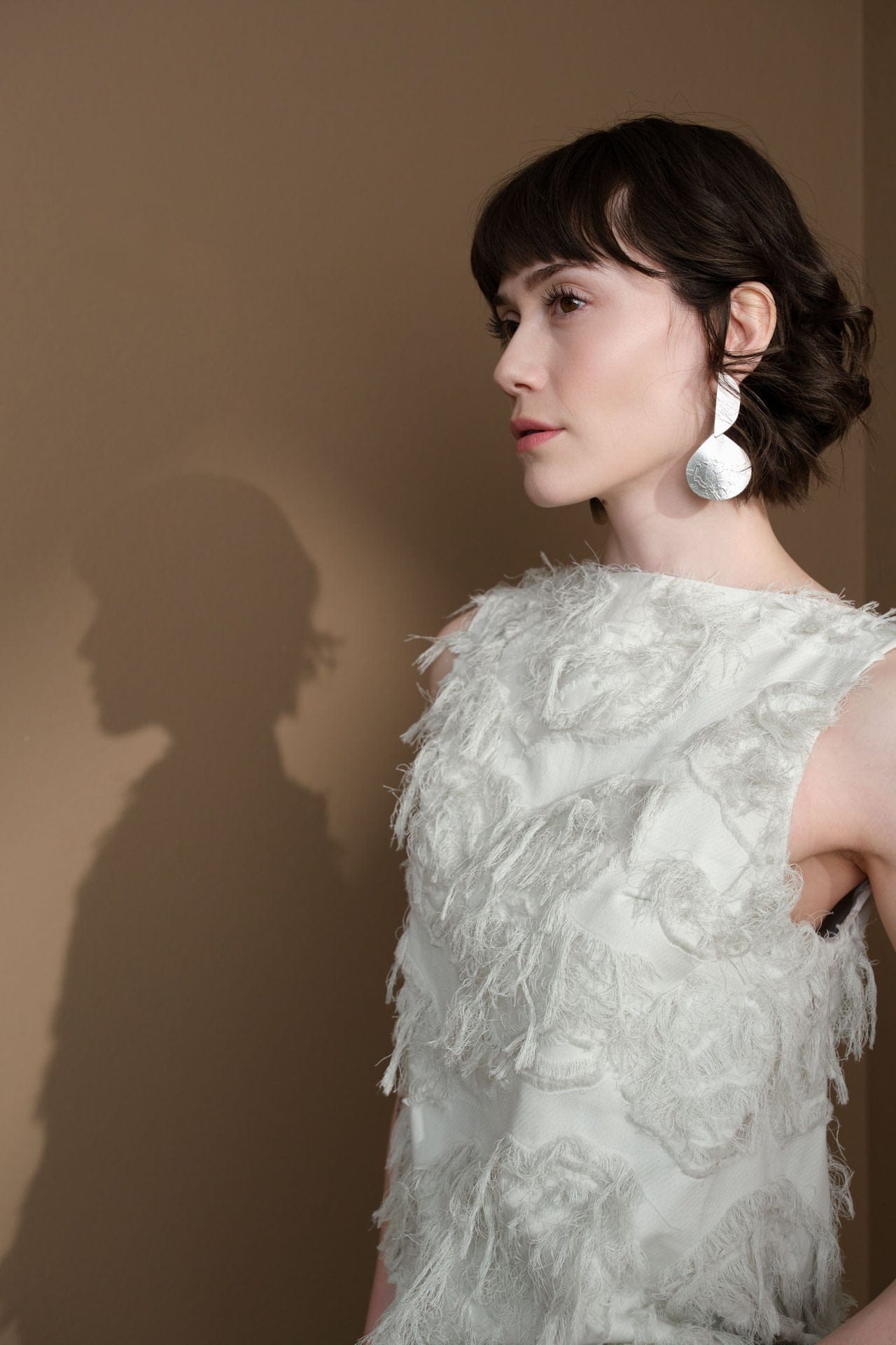 Woman wearing a  Annie Costello Brown Silver Earring with a Studio Nicholson textured white top with feather-like details against a brown background