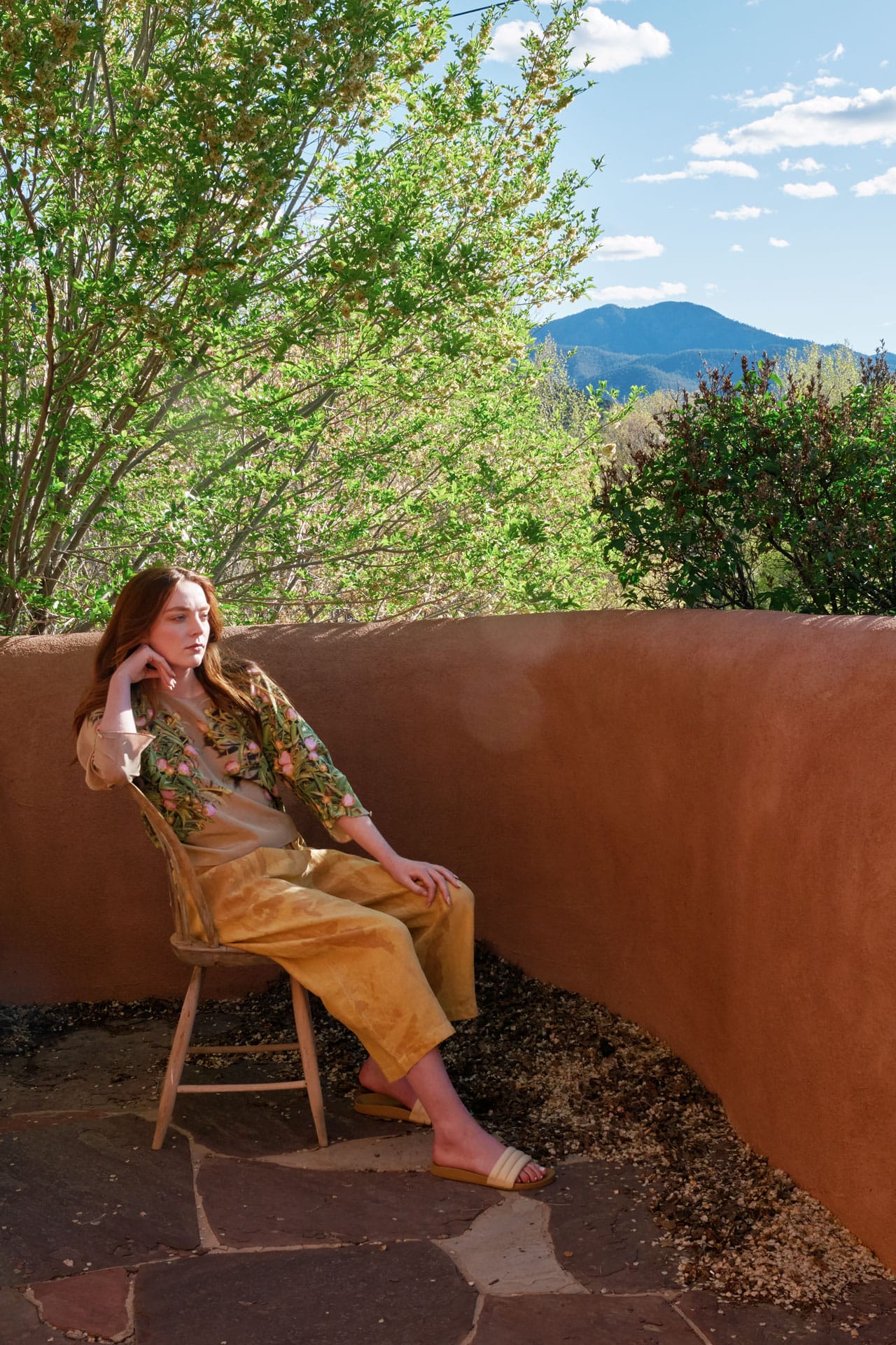 Woman wearing Anntian clothing sitting on a chair against an adobe wall with trees and mountains in the background