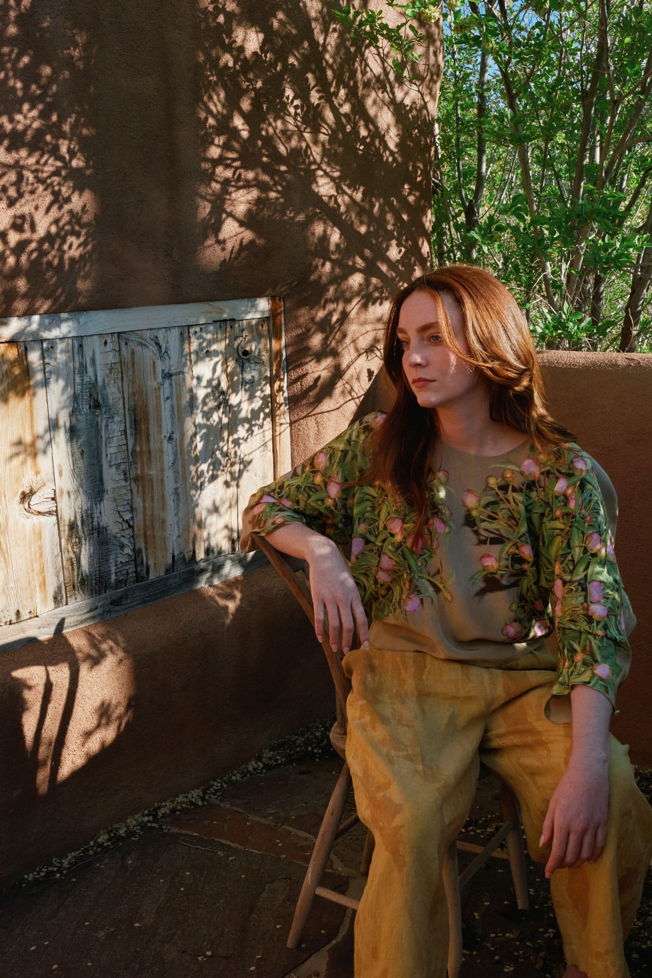 Woman wearing silk foral top from Anntian sitting outdoors on a wooden chair with a rustic wall and greenery in the background