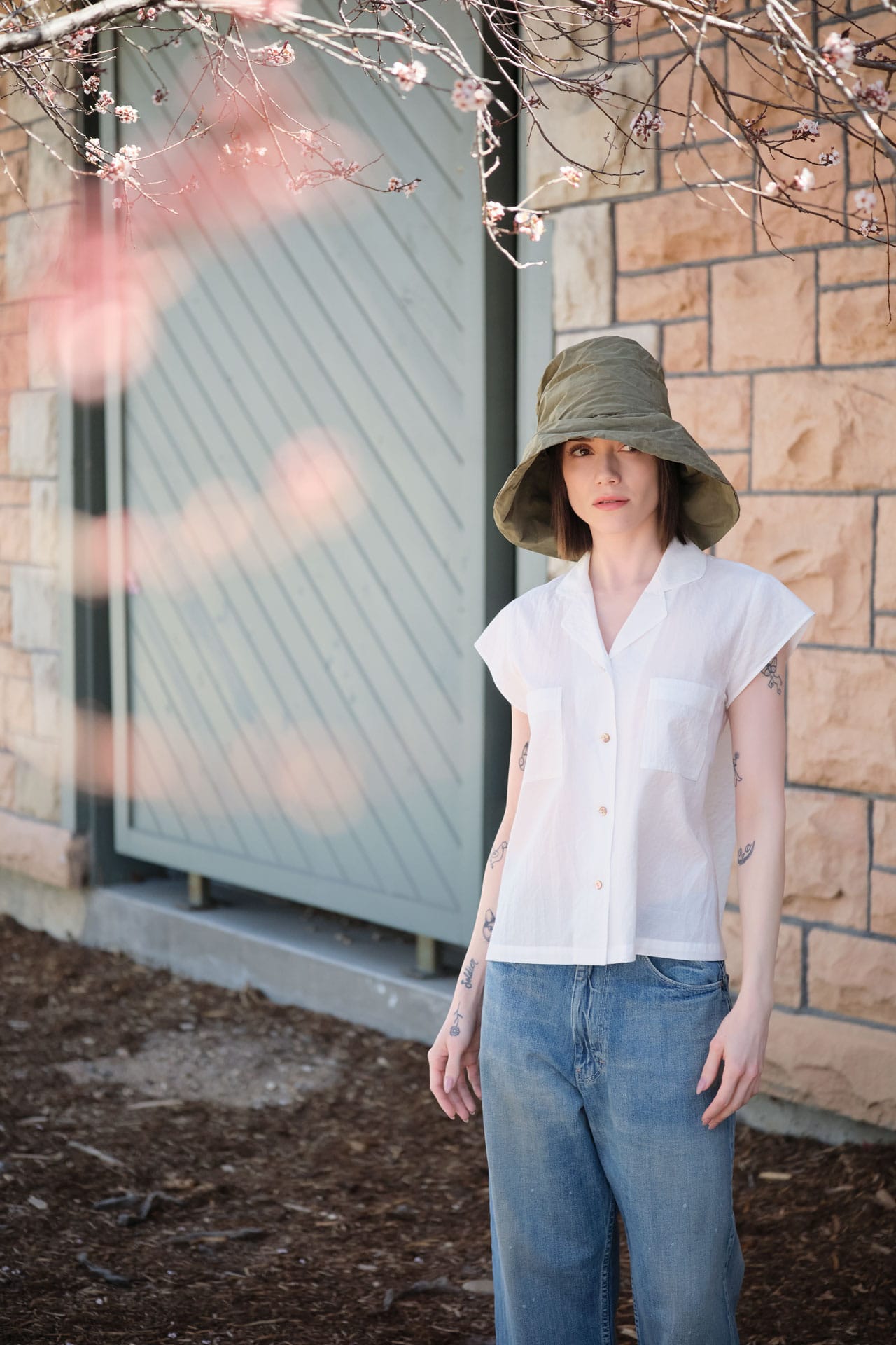 Person wearing a Chimala white shirt and japanese blue jeans with a Reinhard Plank green hat, standing in front of a stone wall and door.