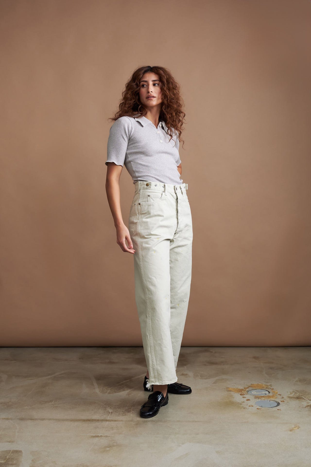 Woman wearing a light gray polo shirt from baserange and white jeans from Chimala against a beige background