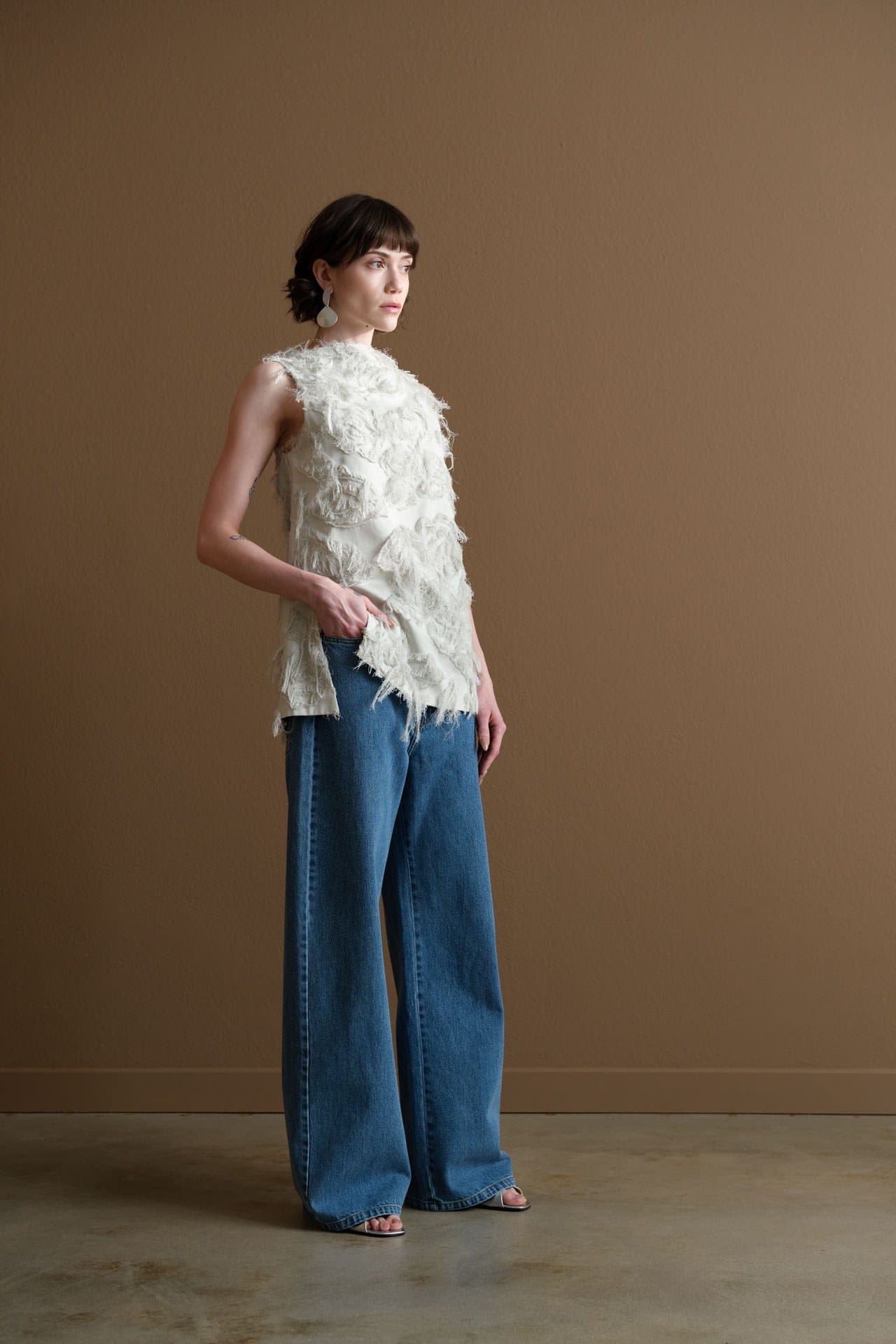 Woman wearing a Studio Nicholson white sleeveless top with ruffled details and Christian Wijnants blue jeans against a brown background.