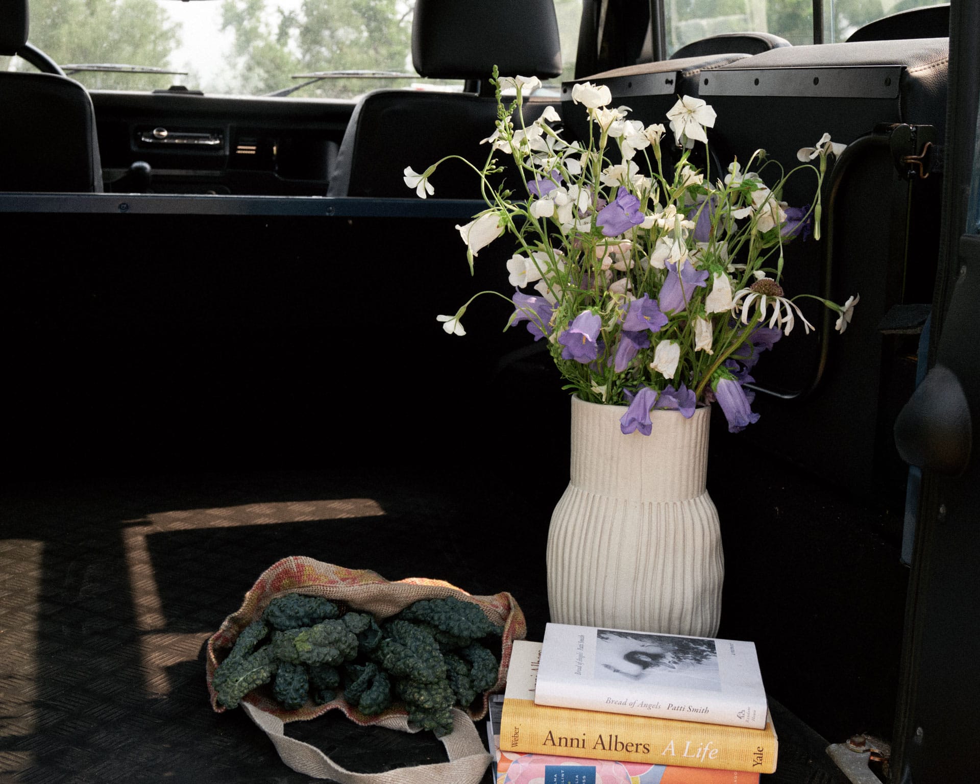 Handmade by CYM Warkov Ceramics, white ribbed ceramic vase filled with white and purple flowers, placed in the back of a dark vehicle next to a canvas tote bag filled with green vegetables and a stack of books.