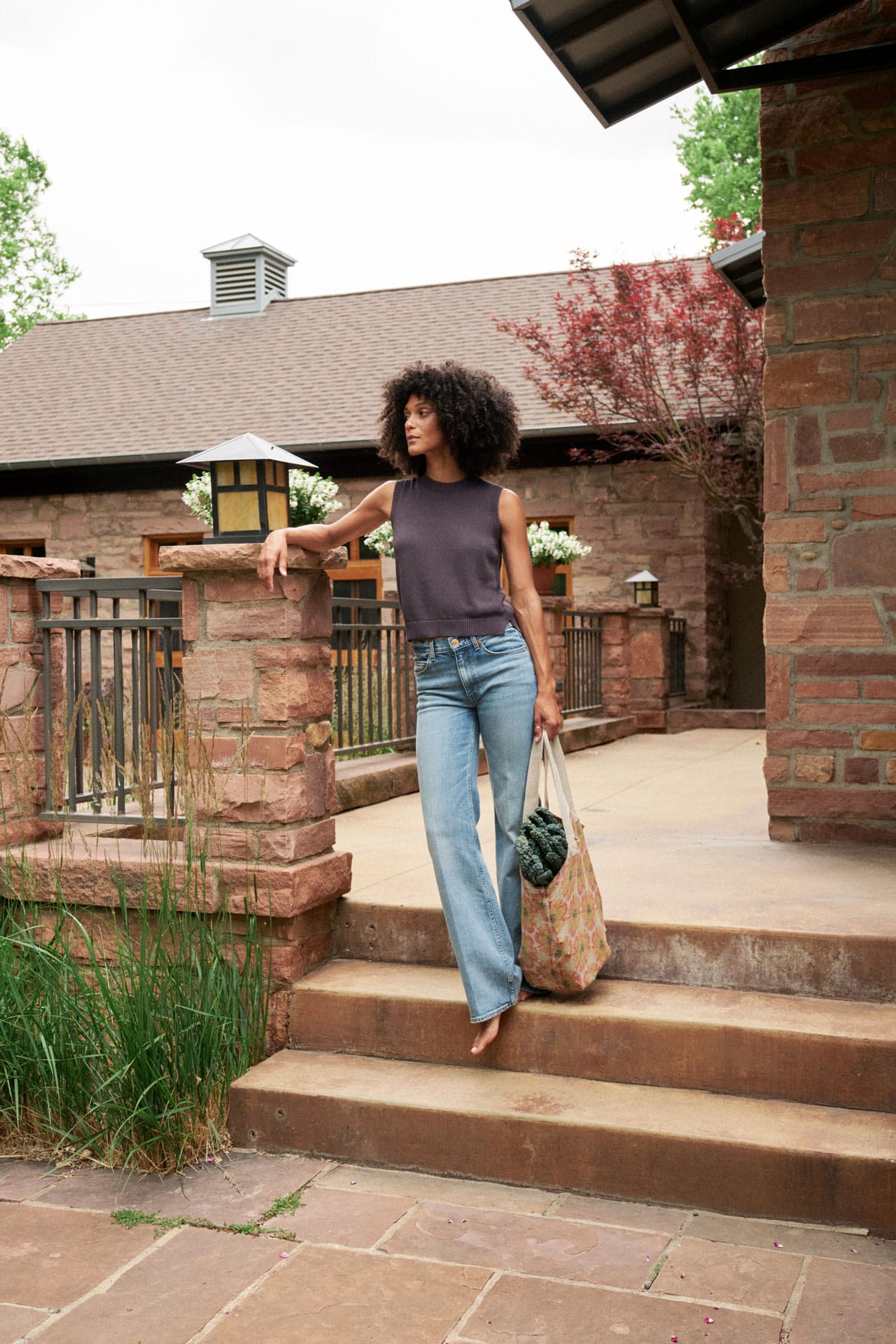 A woman stands on outdoor stone steps in front of a brick house, wearing a sleeveless dark purple knit by Demylee nd light blue flared jeans, holding a woven tote bag.