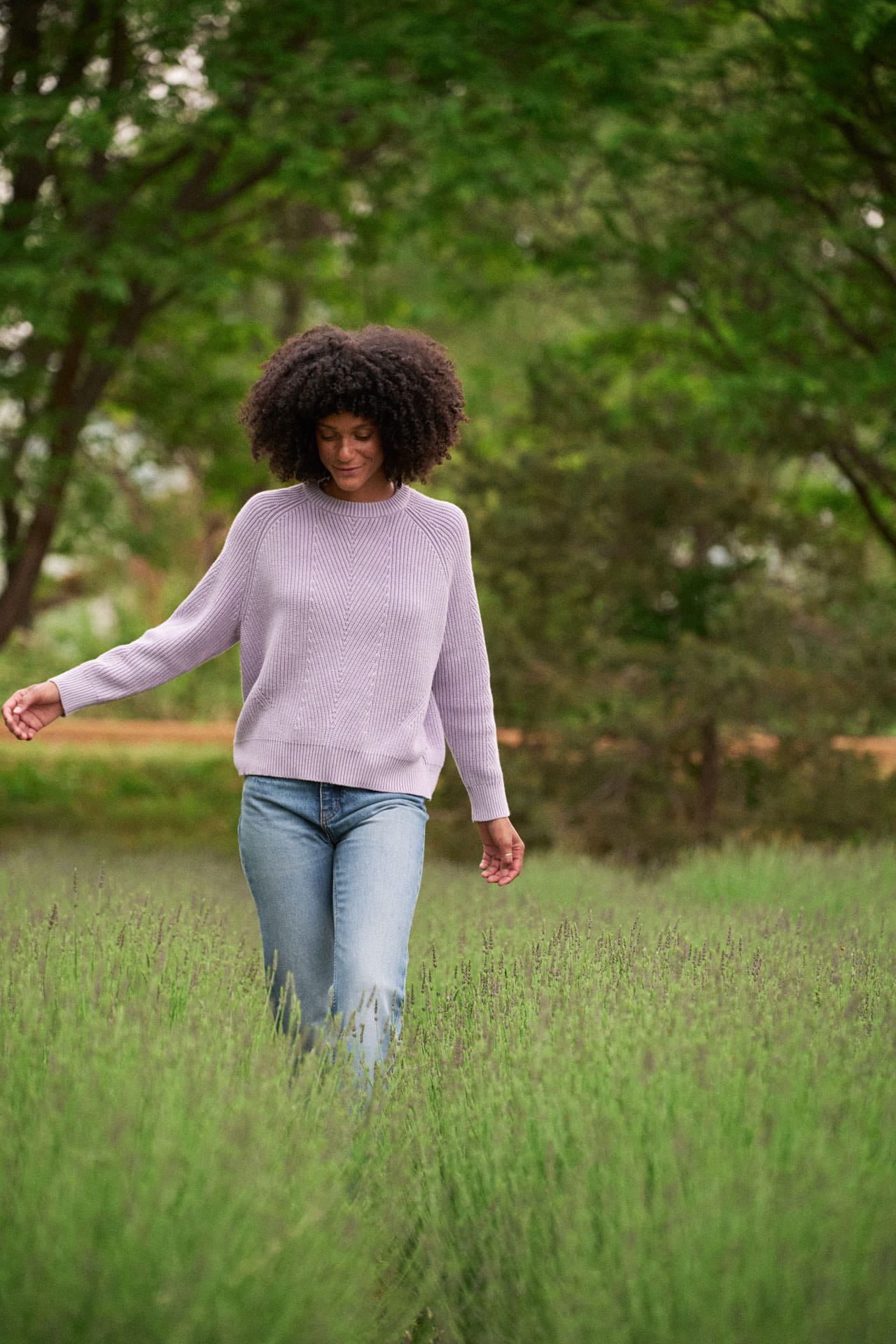 A woman with an afro walking through a green field, wearing a Demylee Chelsea lavender cotton sweater and blue jeans.