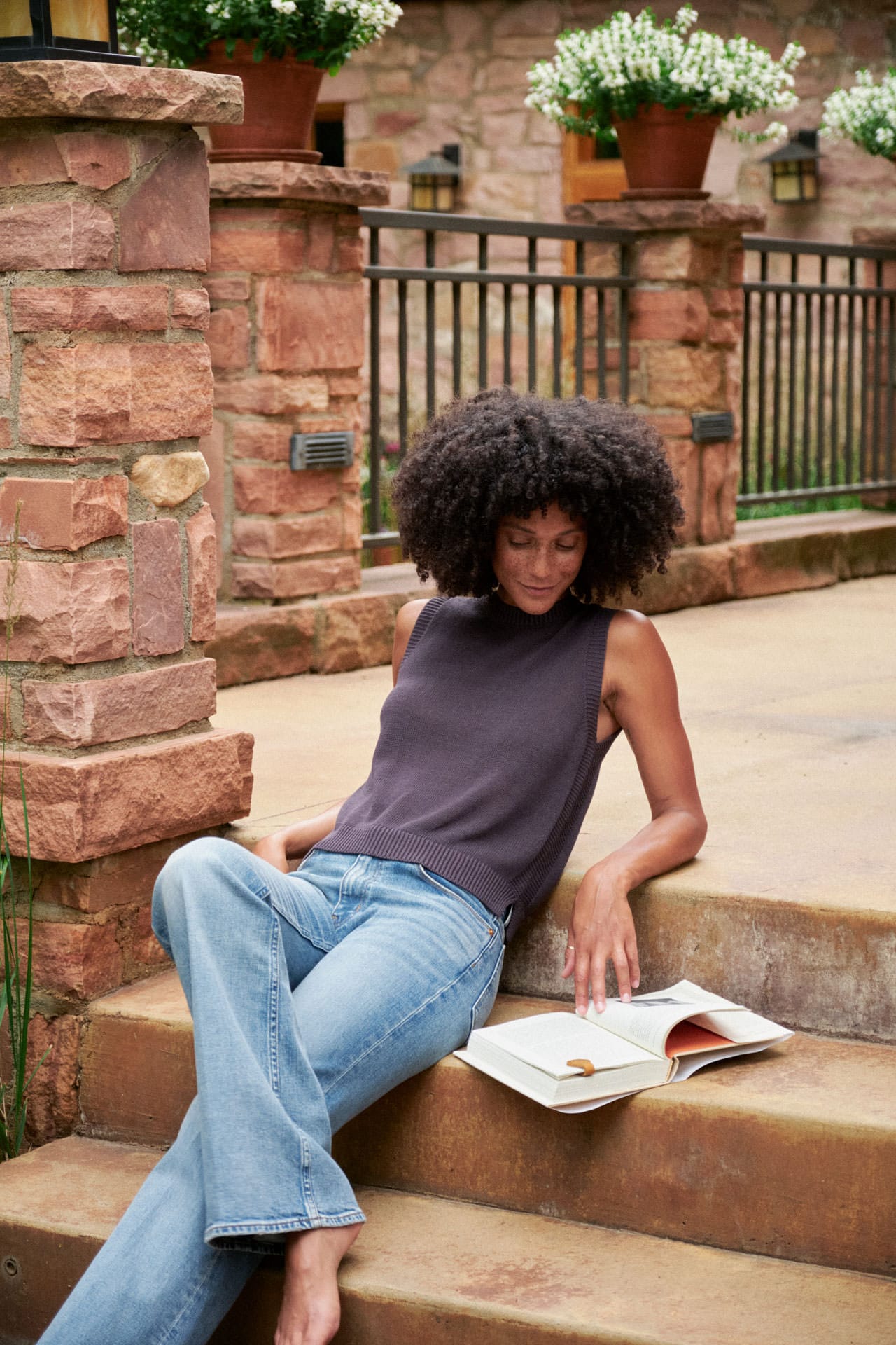A woman sits on stone steps outdoors, wearing a Demylee sleeveless dark gray top and Dala light blue jeans, reading an open book. Stone walls, potted plants, and a railing are visible in the background.