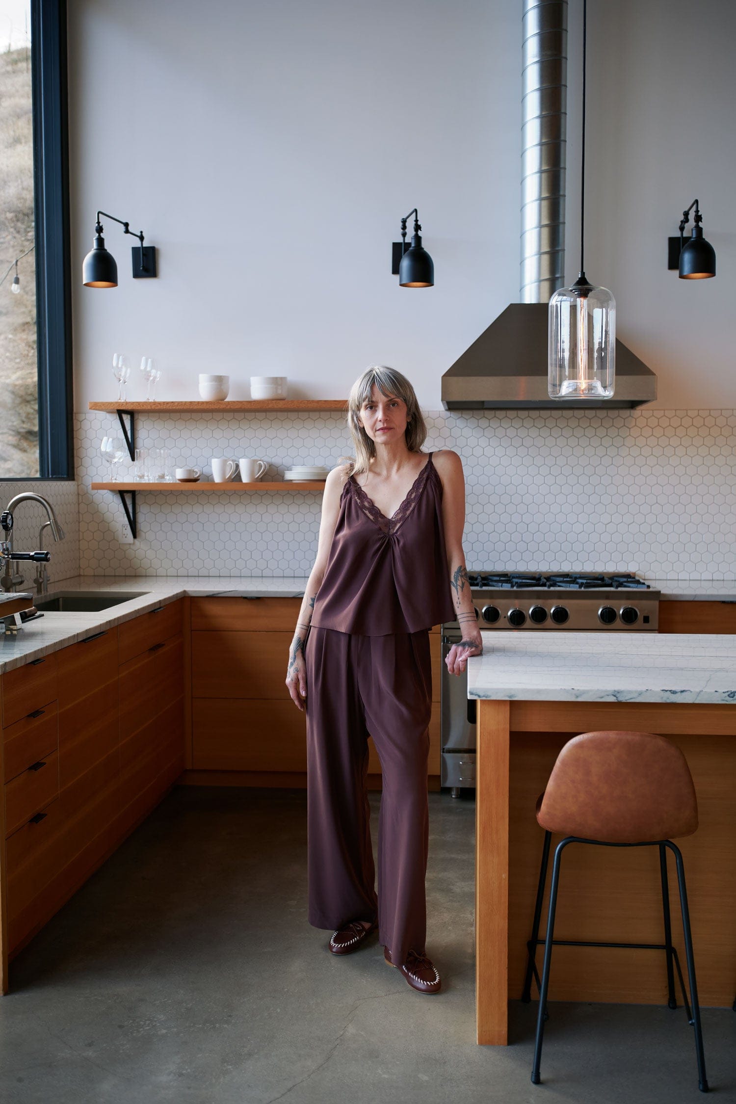 Woman standing in a modern kitchen wearing Jamie Haller clothing and camp loafter with wooden cabinets and white countertops.