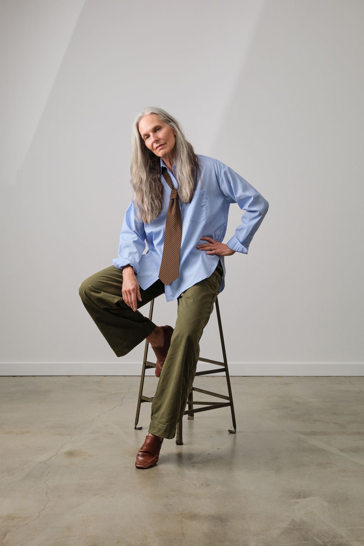 A woman with long gray hair sits on a metal stool in a studio, wearing Jamie Haller's fall collection from head to toe. Features a light blue button-down shirt, olive green wide-leg pants, a brown tie, and brown leather shoes.