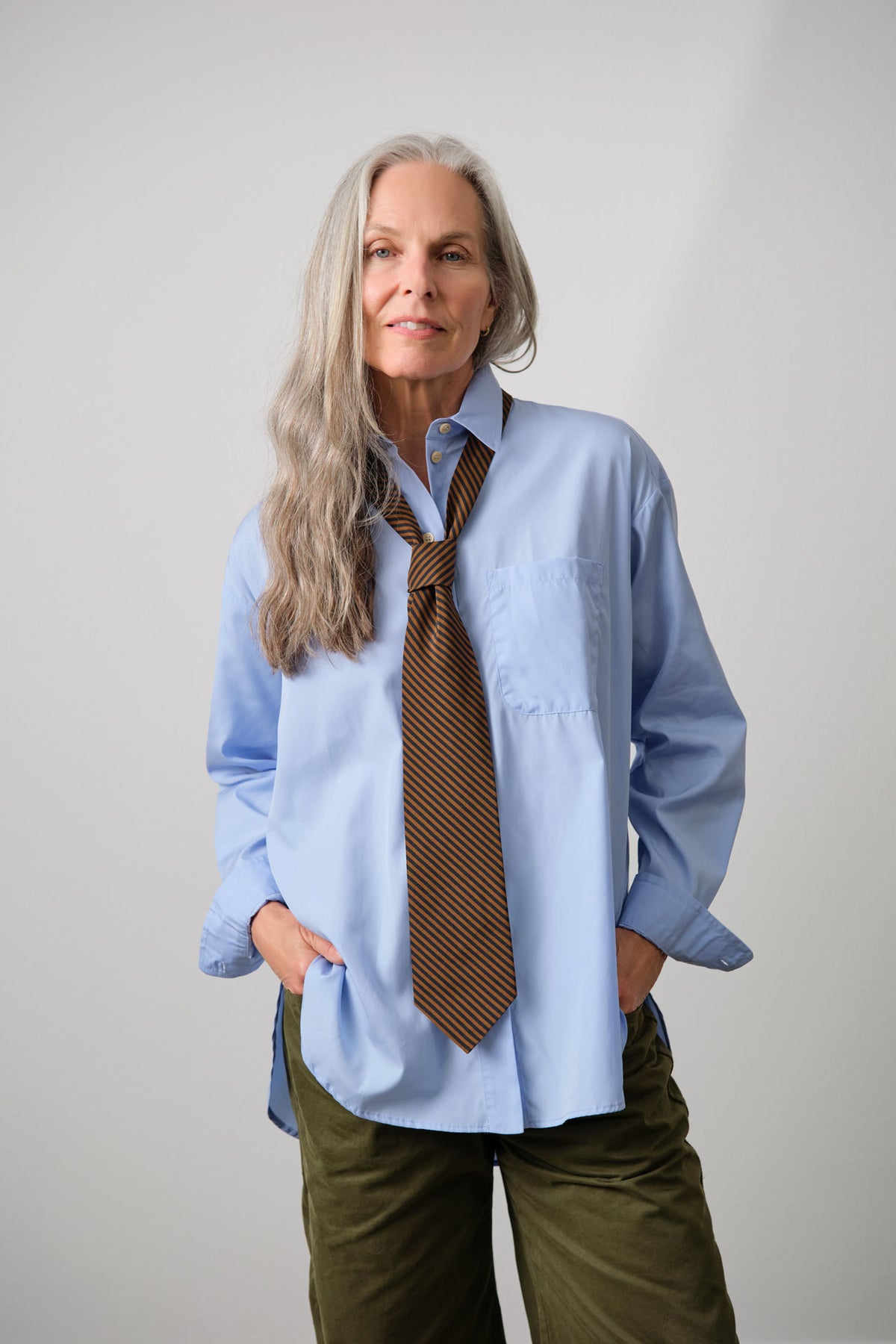 A woman with long gray hair wearing a Jamie Haller light blue button-down shirt, a brown striped necktie, and olive green pants, standing against a light gray studio background.