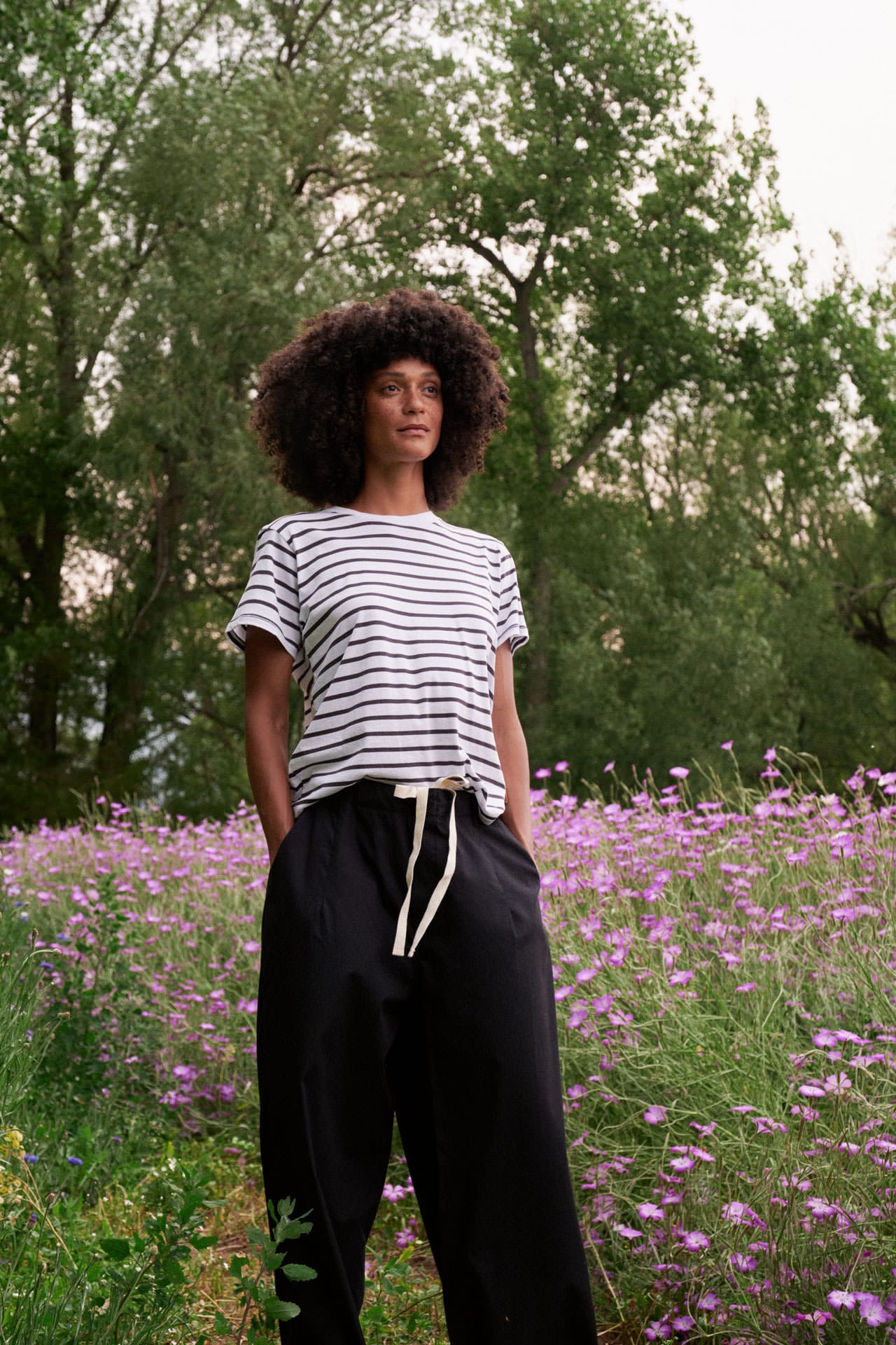 A person standing outdoors in a field of purple flowers, wearing a Jamie Haller white short-sleeve T-shirt with thin black horizontal stripes and loose black drawstring pants.