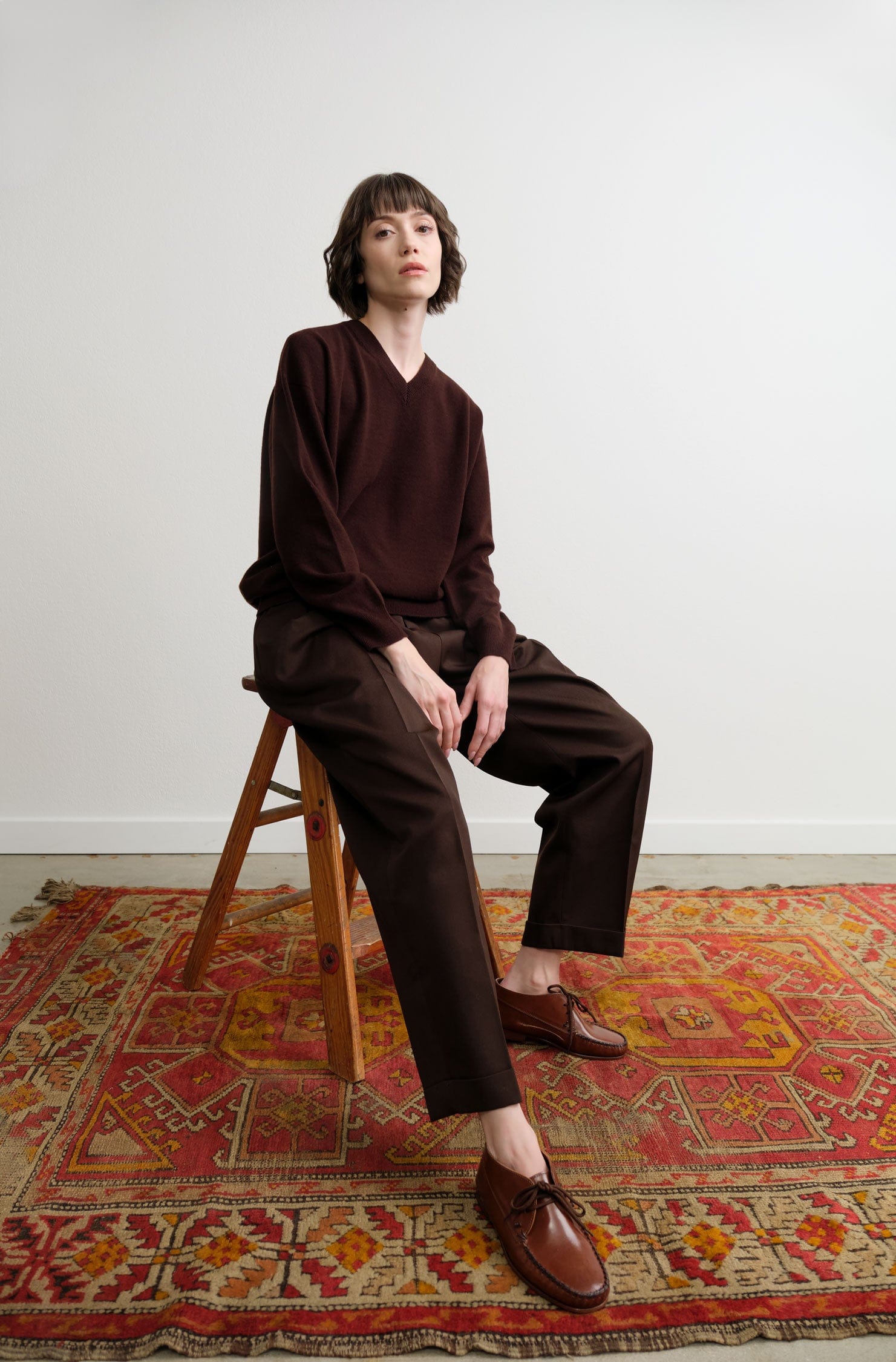 Person wearing a Nicholson & Nicholson brown outfit sitting on a wooden stool on a patterned rug.