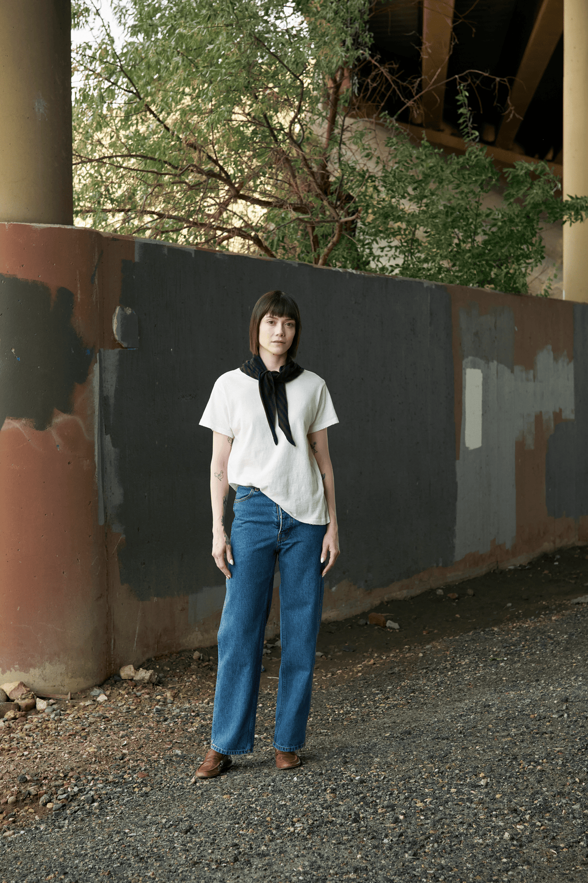 A model stands outdoors under a bridge wearing a Jesse Kamm white short-sleeve top, a black neck scarf, wide-leg blue jeans, and brown shoes, set against a weathered concrete wall and gravel ground.