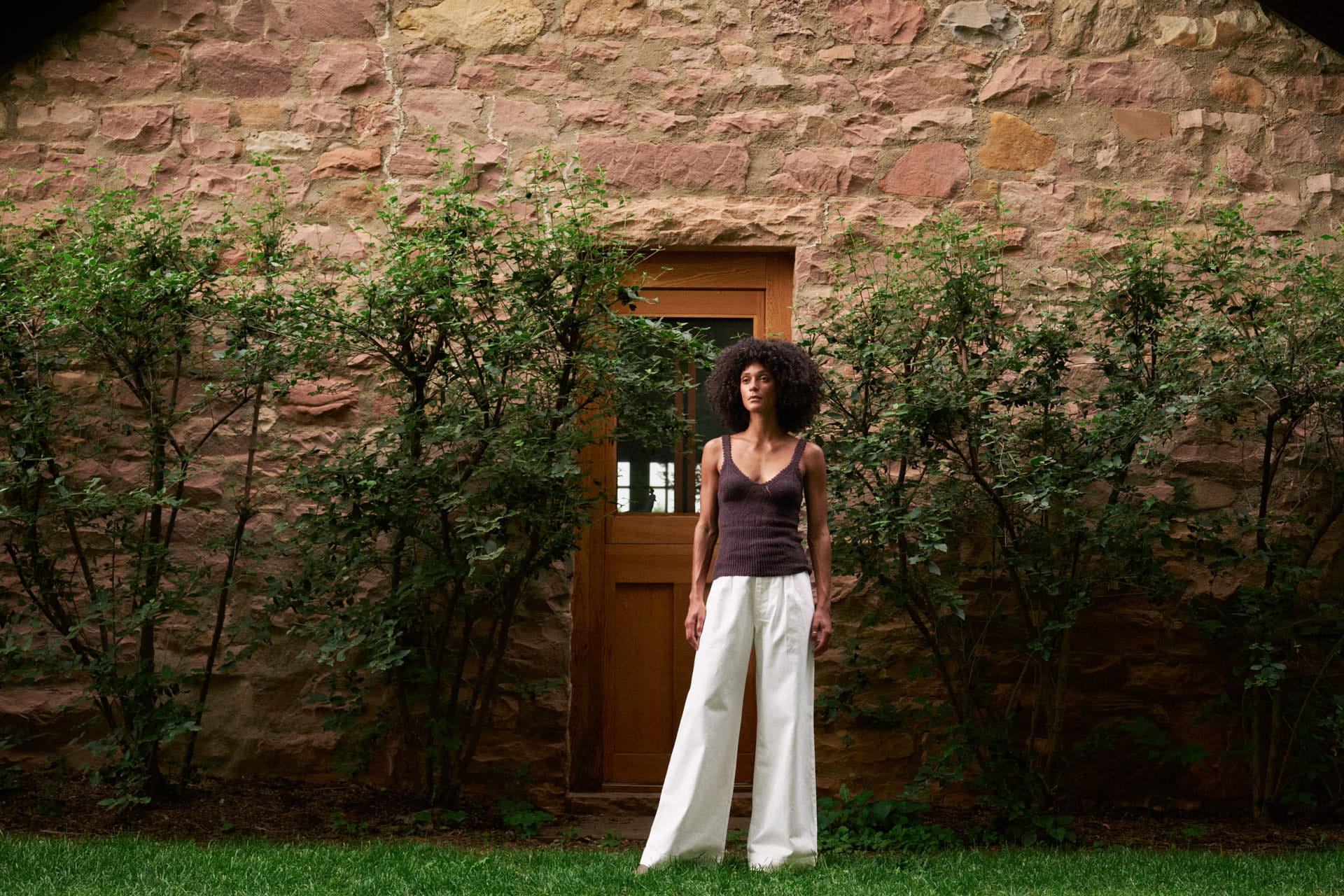 A woman stands outdoors in front of a rustic stone wall and wooden door, wearing a dark brown sleeveless top by rachel comey and jesse kamm wide-leg white pants.