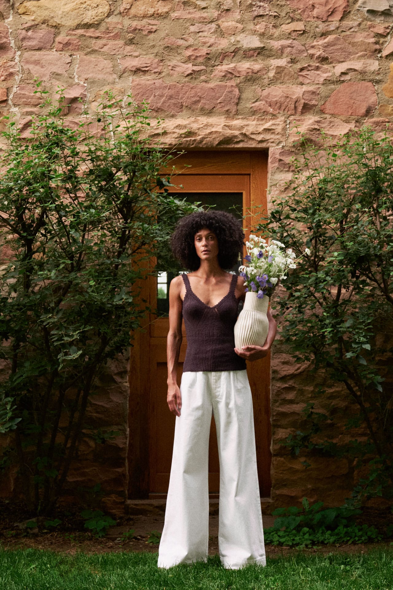 A woman in front of a rustic stone wall and wooden door, wearing a rachel comey dark brown sleeveless top and jesse kamm white wide-leg pants while holding a white ceramic vase with flowers.