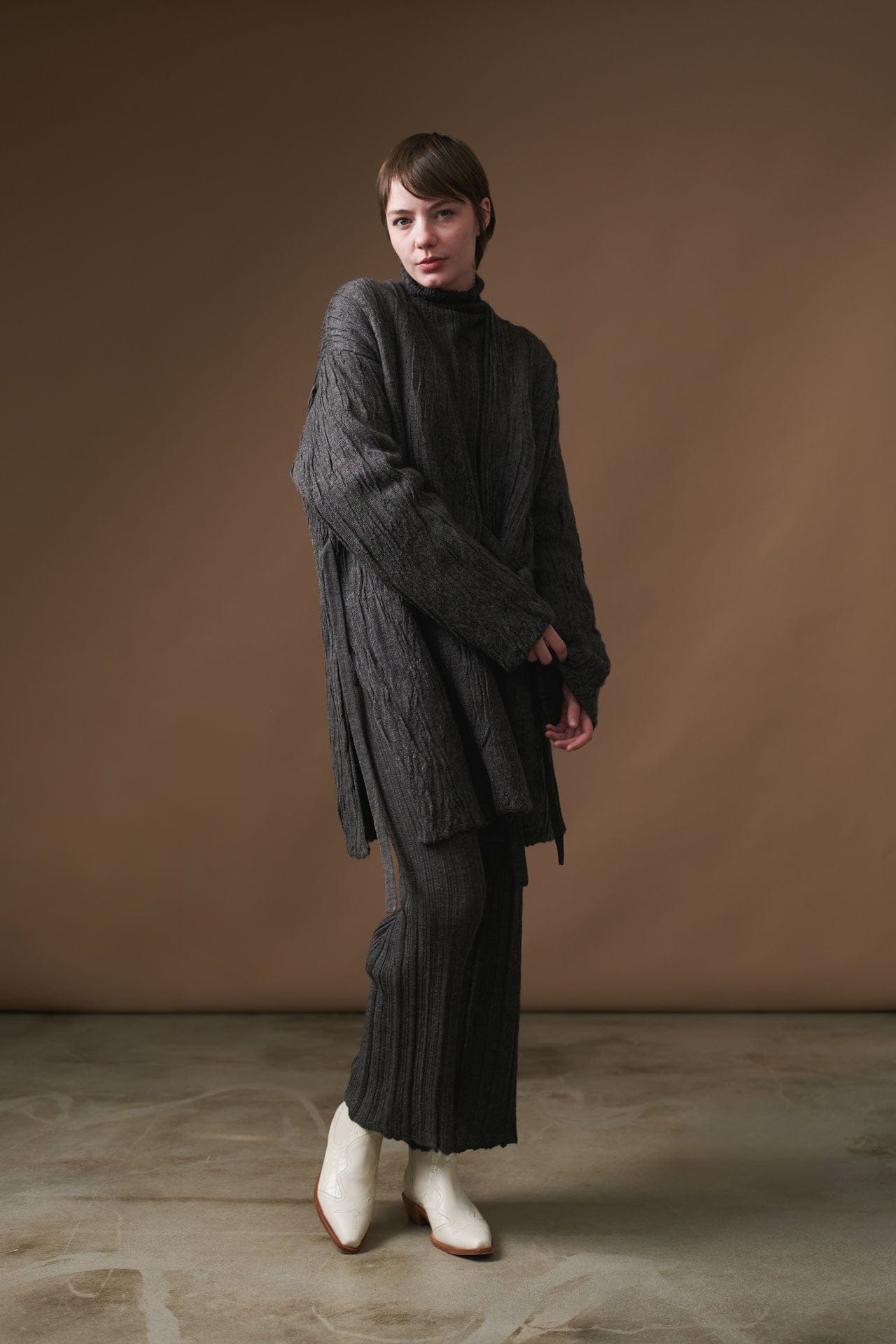 Person wearing a Lauren Manoogian dark textured outfit with white boots against a brown background