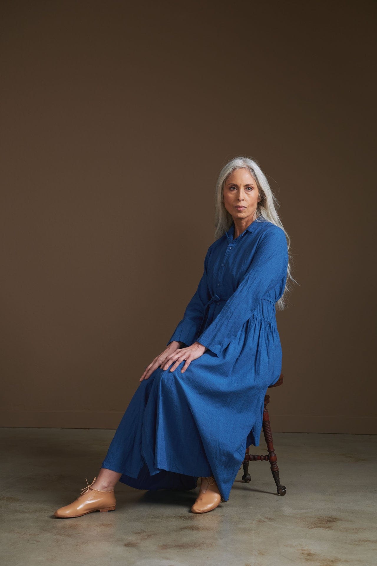Woman wearing a blue pas de calais dress sitting on a stool against a brown background