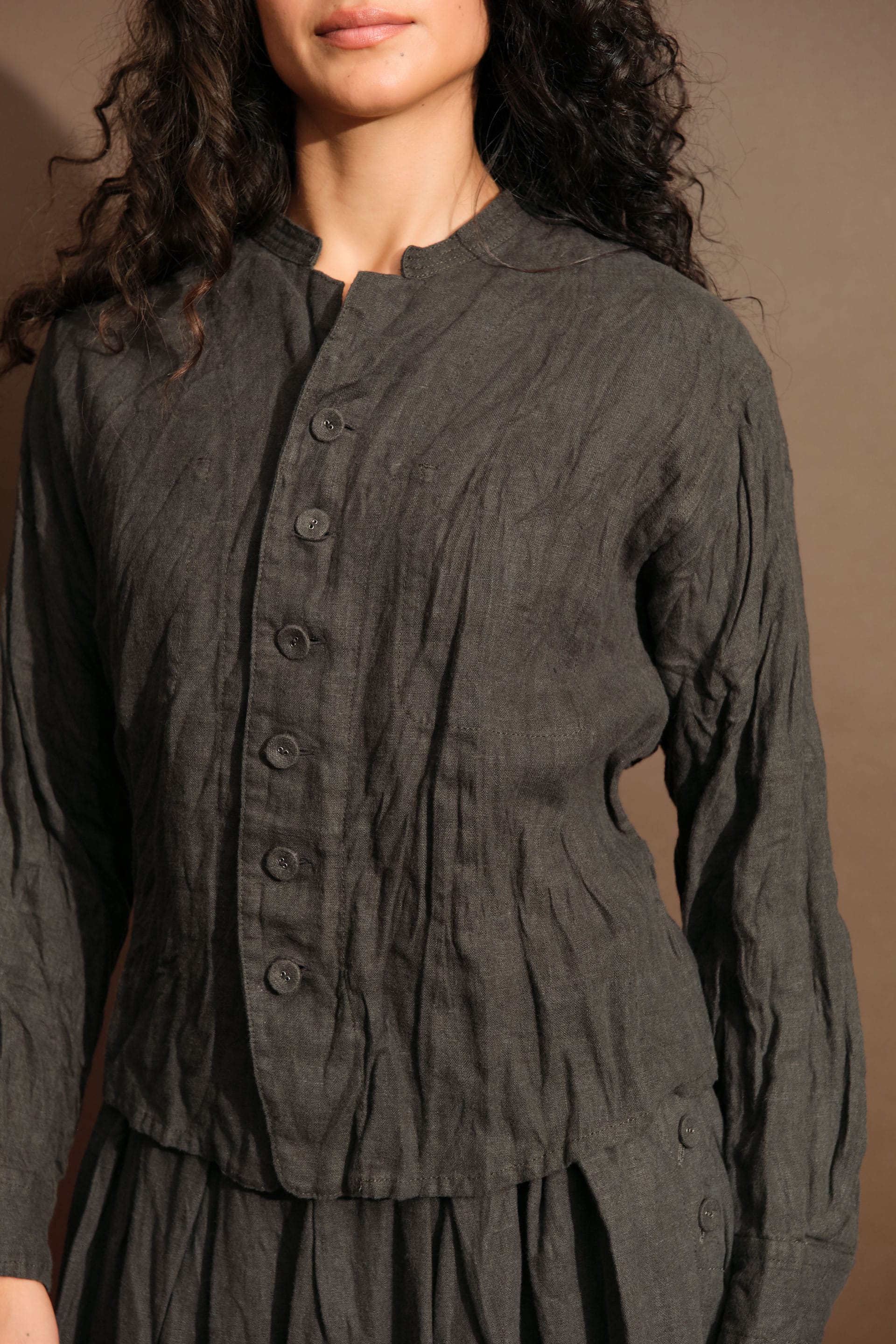 A close-up of a woman wearing a dark charcoal gray crinkled linen button-up blouse from Pas de Calais with a high collar and long sleeves, set against a warm brown background.