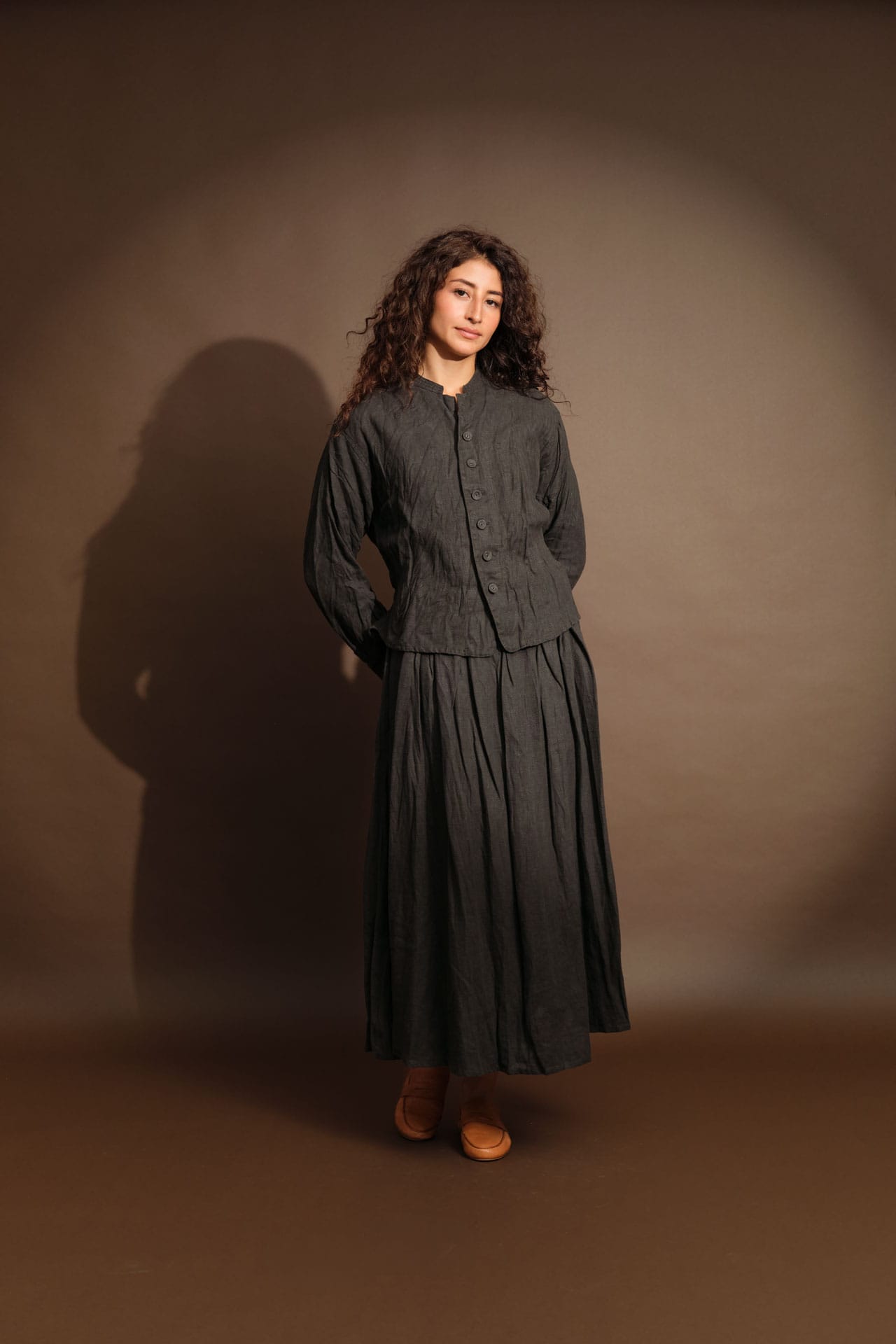 A woman wearing new Pas de Calais stands against a warm brown studio backdrop. She is wearing Jamie Haller shoes, and her shadow is cast on the wall behind her.