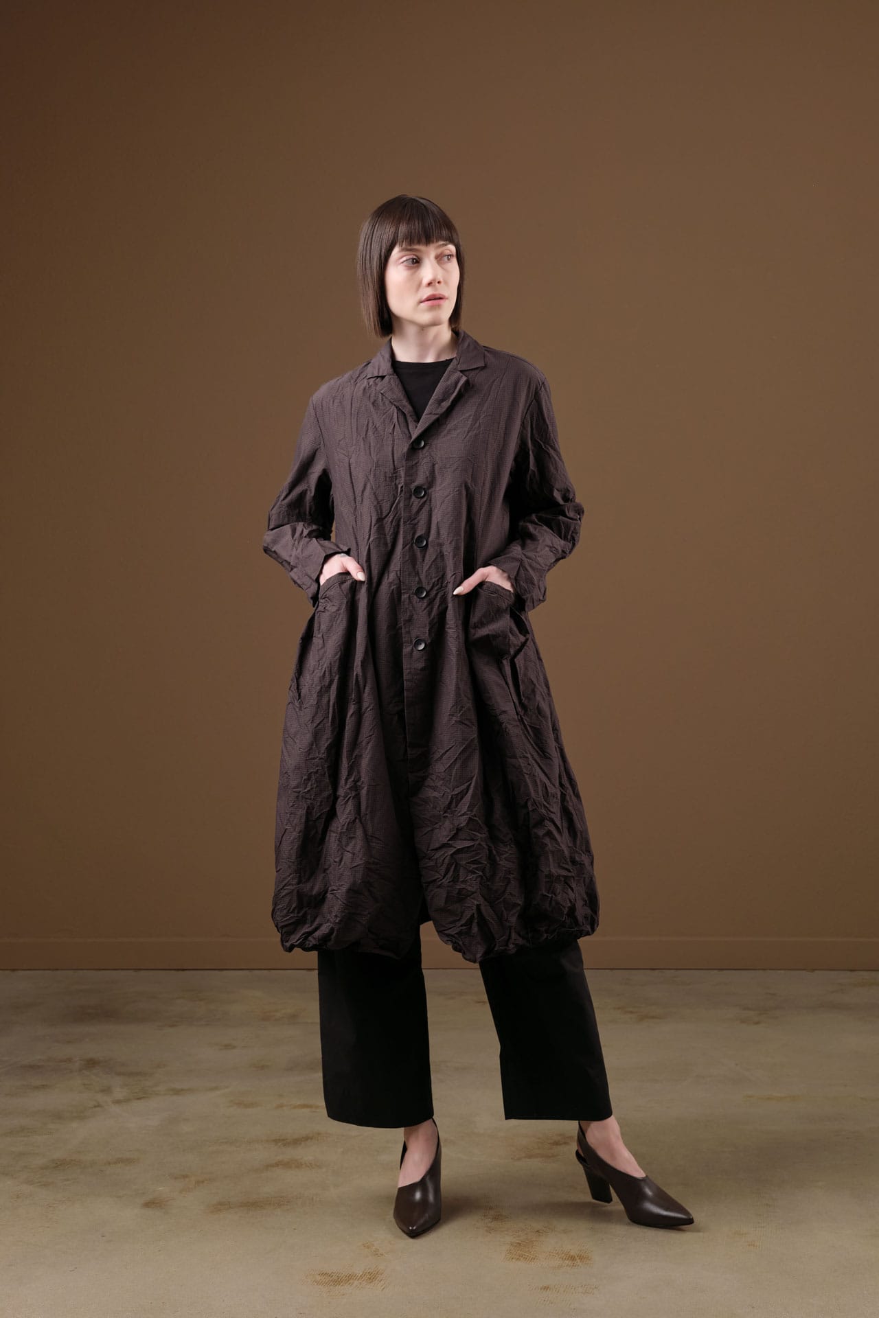 A model stands against a solid brown studio backdrop wearing a dark brown crinkled, oversized long coat by Pas de calais over a black outfit with black wide-leg pants and dark brown heels.