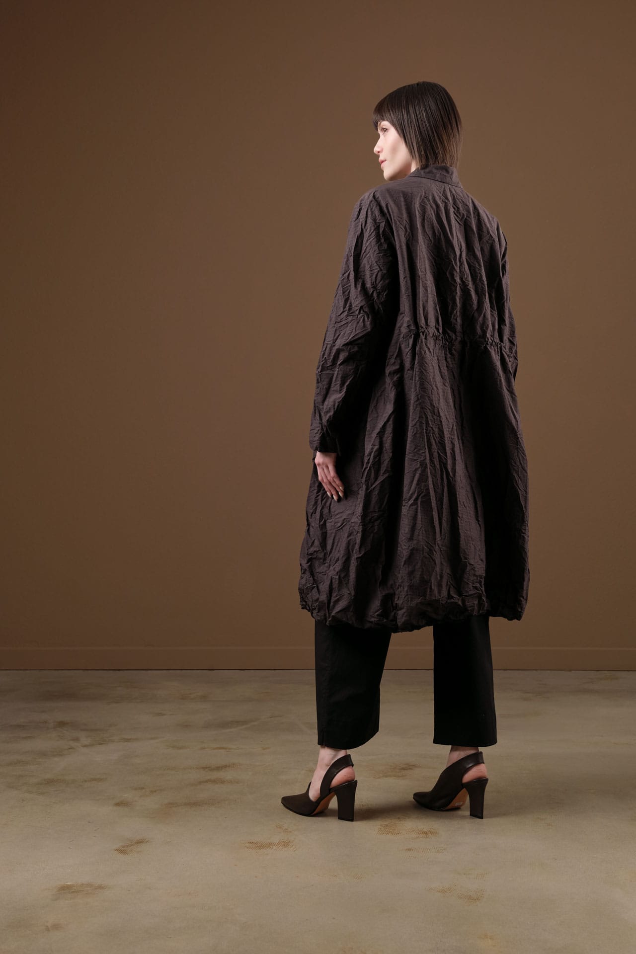 A model is shown from the back wearing a dark brown crinkled, textured long coat by pas de calais over black pants and black high-heeled sandals, standing against a solid brown studio backdrop.