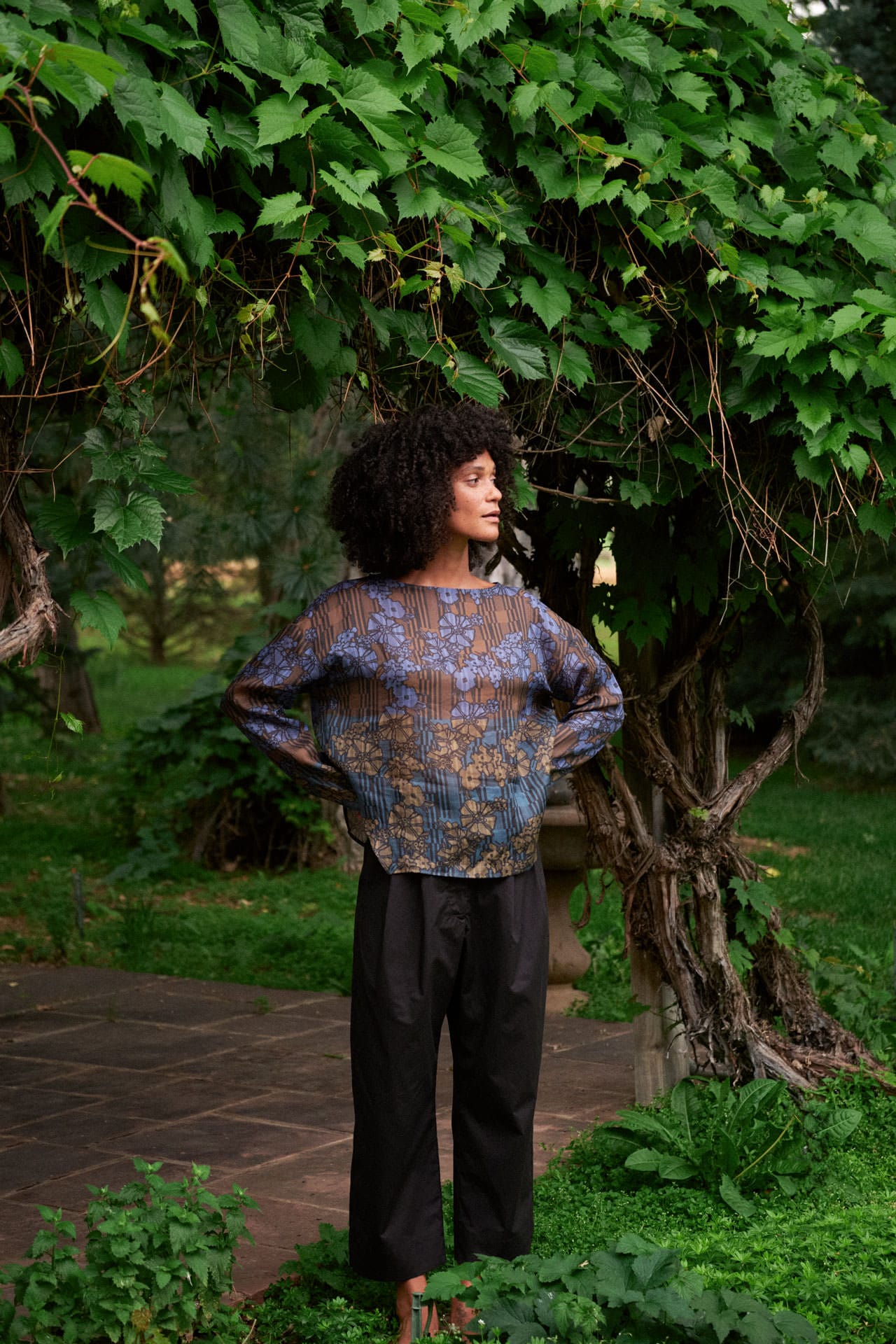 A woman with curly black hair stands outdoors in a garden under a leafy tree, wearing a rachel comey floral barter top long-sleeve top with blue, purple, and gold tones, paired with black pants.