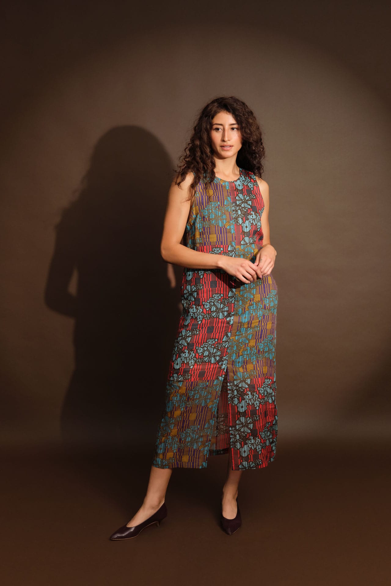 A woman modeling a sleeveless midi dress by Rachel Comey with a colorful abstract pattern in red, blue, green, and yellow, standing against a dark brown studio backdrop with a soft shadow.