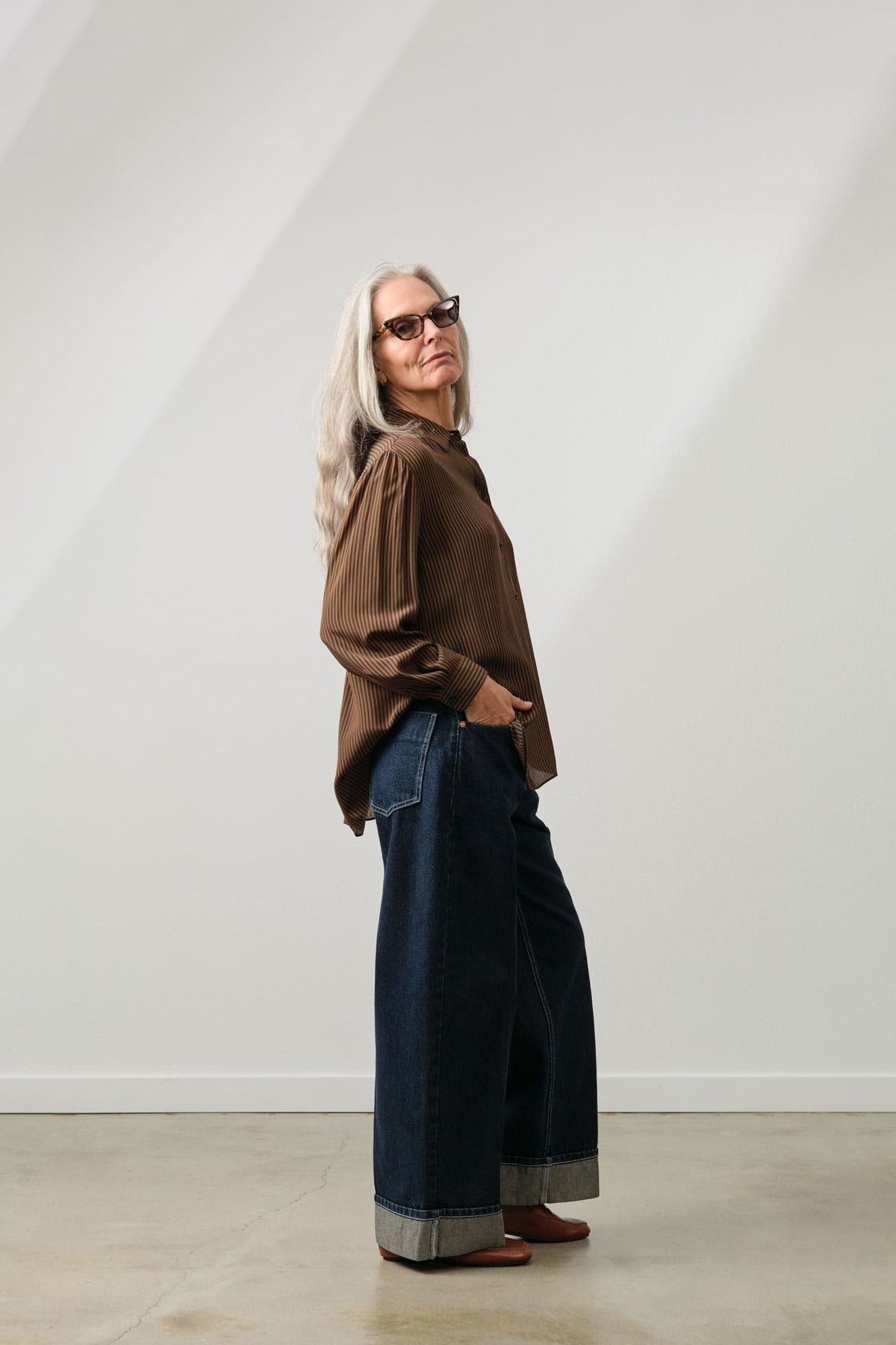 A woman with long gray hair and sunglasses stands in profile wearing a Jamie Haller brown long-sleeve blouse and Rachel Comey wide-leg dark blue jeans with rolled cuffs, against a minimalist white wall with soft diagonal shadows.
