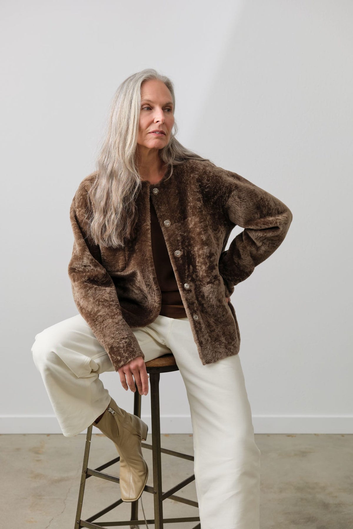 A woman with long gray hair sits on a metal stool wearing a Rachel Comey Shearling brown textured fuzzy button-up jacket, a dark brown top, cream wide-leg pants, and tan ankle boots against a light gray wall.