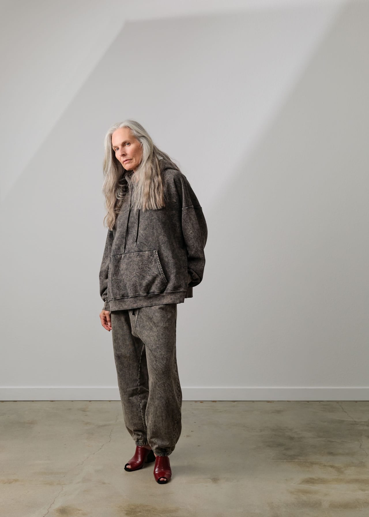A person with long gray hair stands in a minimalist studio wearing a Rachel Comere dark gray marled revere hoodie and matching jogger link pants, paired with dark red boots.