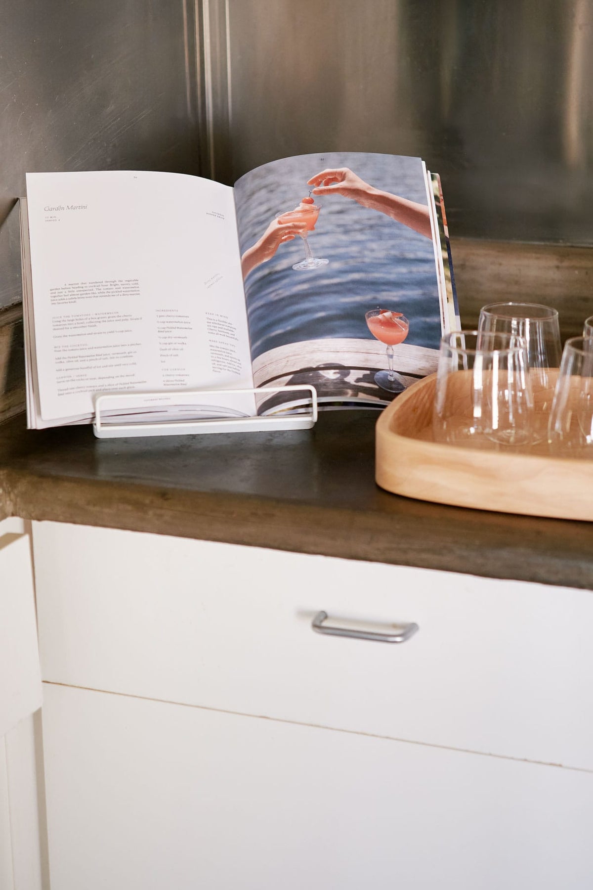 Through Sunday recipe magazine displayed on a dark countertop next to a wooden tray holding clear malfati drinking glasses, with white cabinetry below.