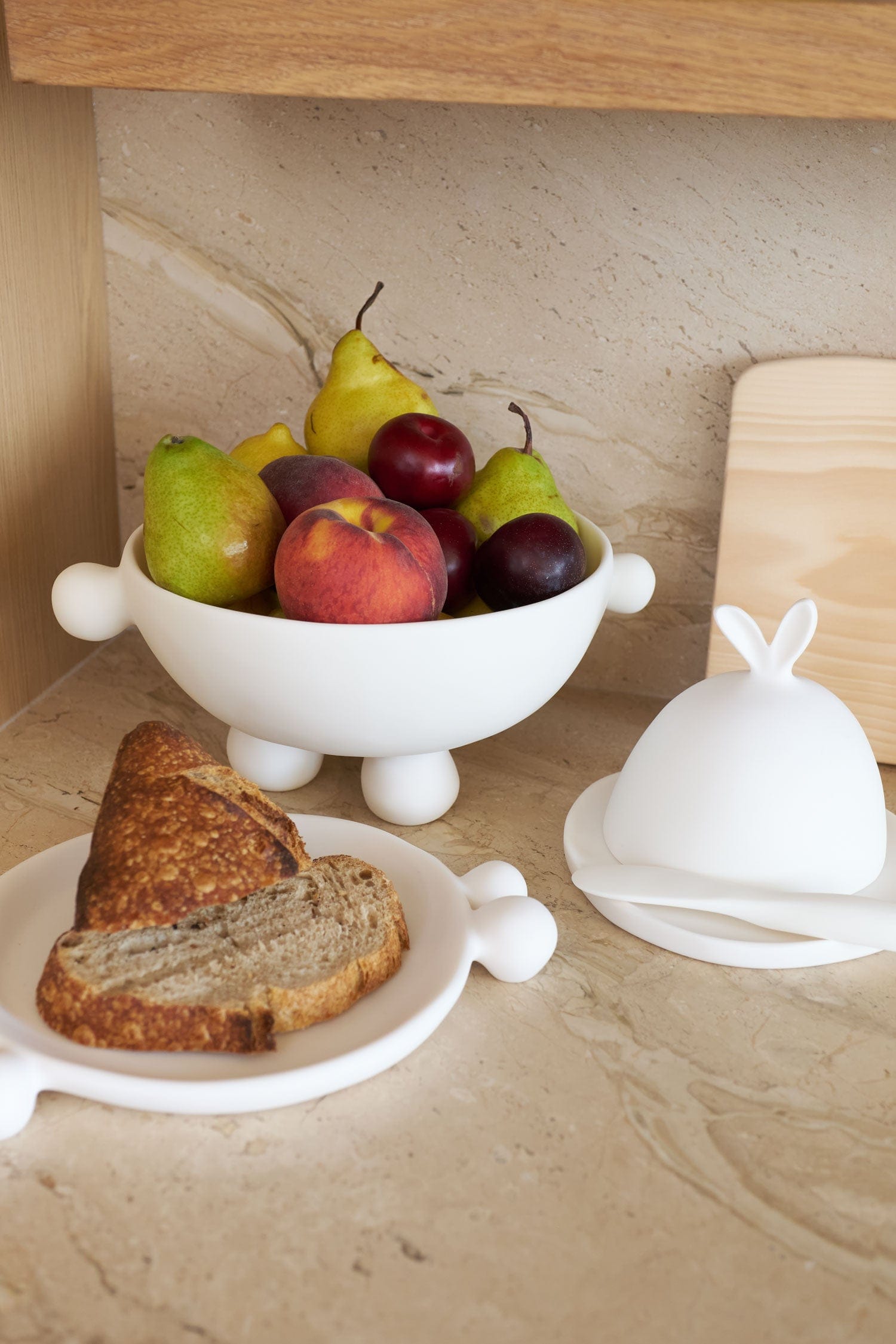 Fruit bowl from Tina Frey Designs with pears, apples, and a peach on a kitchen counter with bread and a white container.
