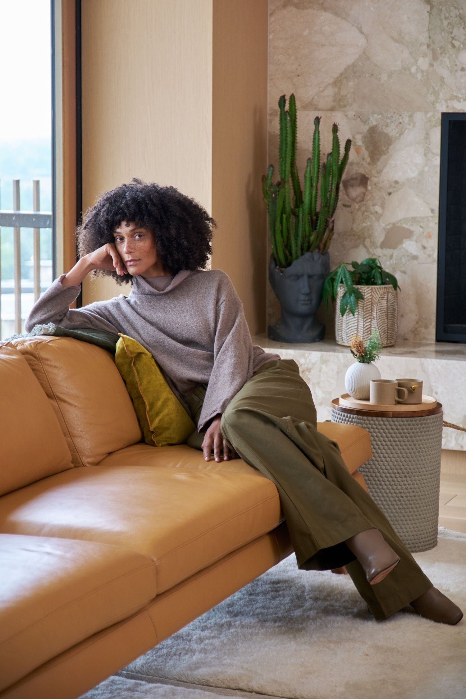 Woman sitting on a brown leather sofa wearing an Elsa Esturgie sweater and Christian Wijnants pants in a modern living room.
